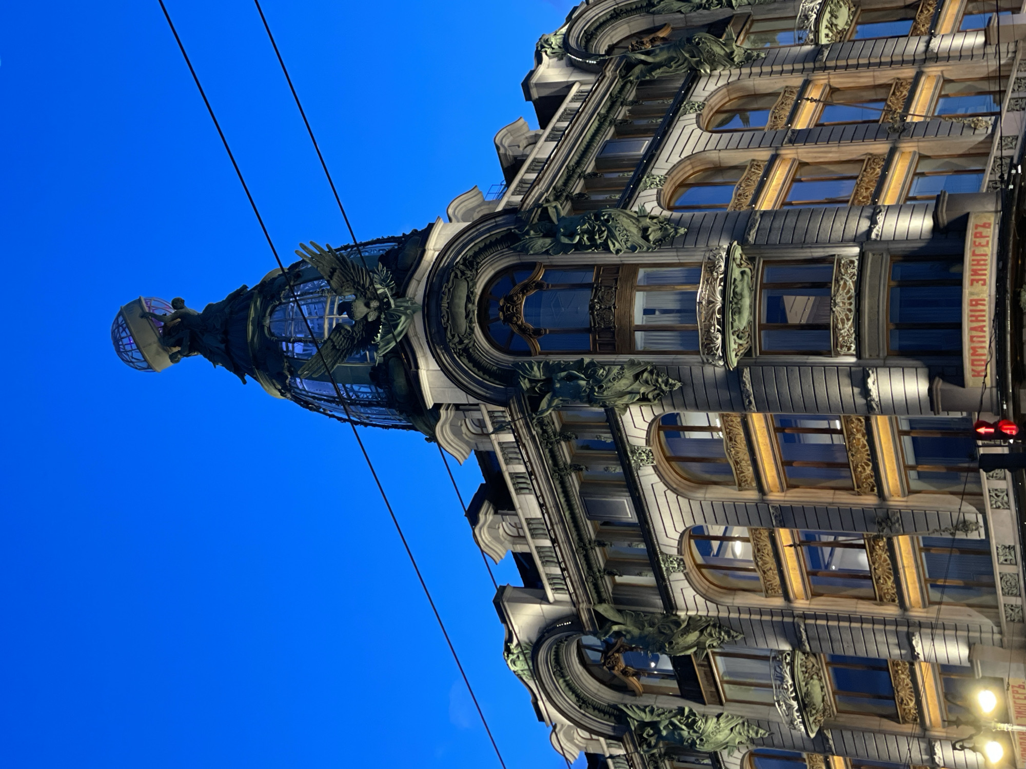 The image displays a grand, multi-story building rendered in an ornate architectural style, likely Art Nouveau, viewed from a low angle at twilight or night. The deep blue sky above indicates the time of day, with artificial lights illuminating the building's intricate facade.
A prominent feature is the elaborate glass dome or cupola at the top of the building, which is crowned by a large globe supported by a sculptural figure. Large, detailed eagles with outstretched wings flank the dome. The building's exterior is rich with decorative elements, including ornate window frames, balconies, and intricate relief work. The windows vary in shape, with several arched and rectangular designs visible, reflecting the ambient light.
The facade is well-lit, highlighting the architectural details and creating a striking contrast against the dark sky. Overhead electrical wires are visible stretching across the upper portion of the frame. On the far right side of the building, vertically oriented text in Cyrillic reads "КОМПАНИЯ ЗИНГЕРЪ" (Kompaniya Zinger), which translates to "Singer Company." The scene depicts a majestic, historically significant urban structure, characteristic of Saint Petersburg, Russia.