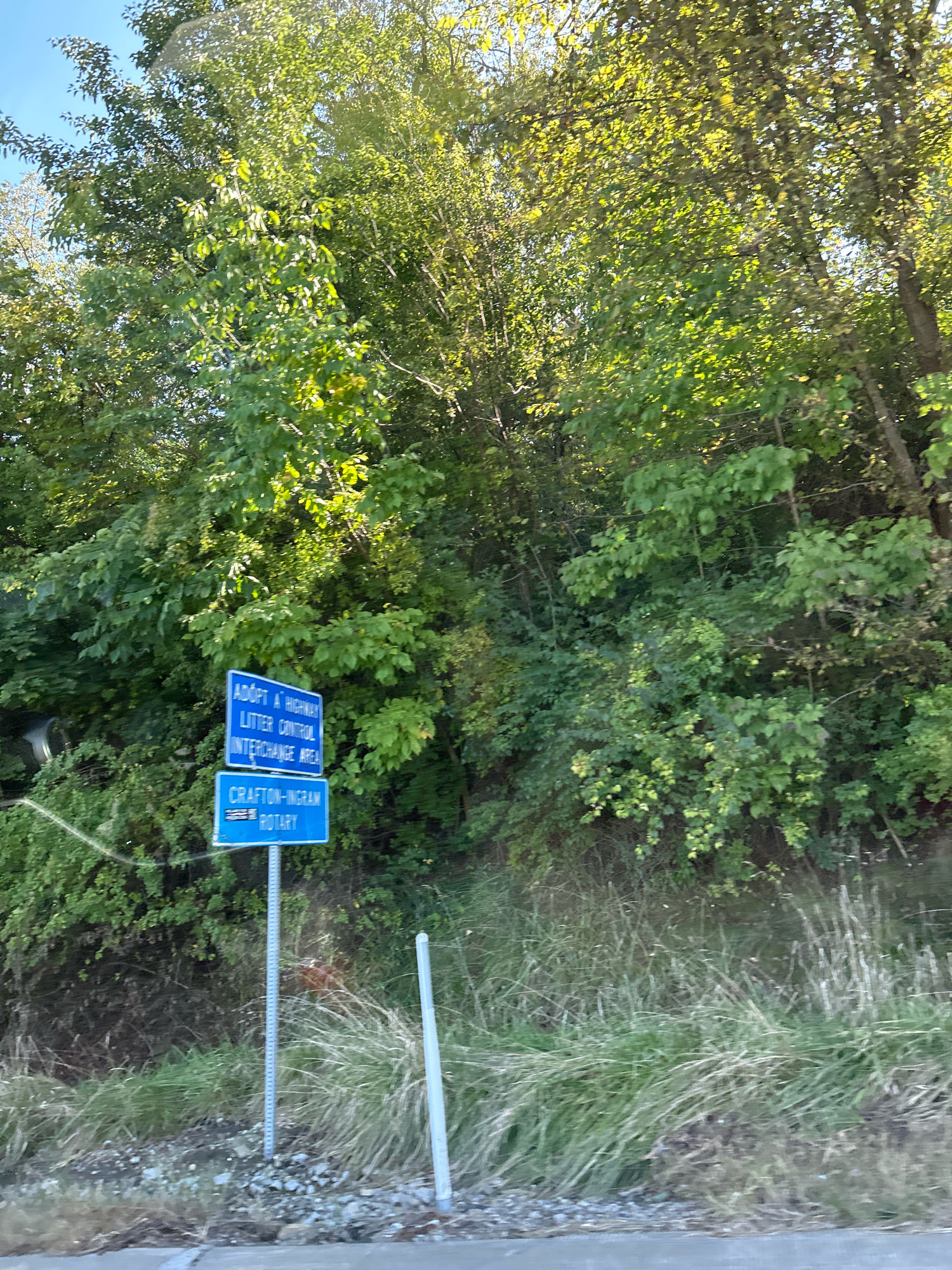 The image shows a roadside scene with two blue signs mounted on metal posts. The upper sign reads "ADOPT A HIGHWAY LITTER CONTROL INTERCHANGE AREA". The lower sign reads "CRAFTON-INGRAM ROTARY". The signs are set against a backdrop of lush green trees and vegetation. Tall, dry grass is visible in the foreground, and the ground appears to be covered with small stones and dirt. In the distance, a bright sky is visible through the canopy of leaves. The scene is set during the daytime, with sunlight filtering through the trees, creating dappled light effects. The overall impression is one of a well-maintained roadside area with natural surroundings. The location is identified as being in Robinson Township, United States of America.