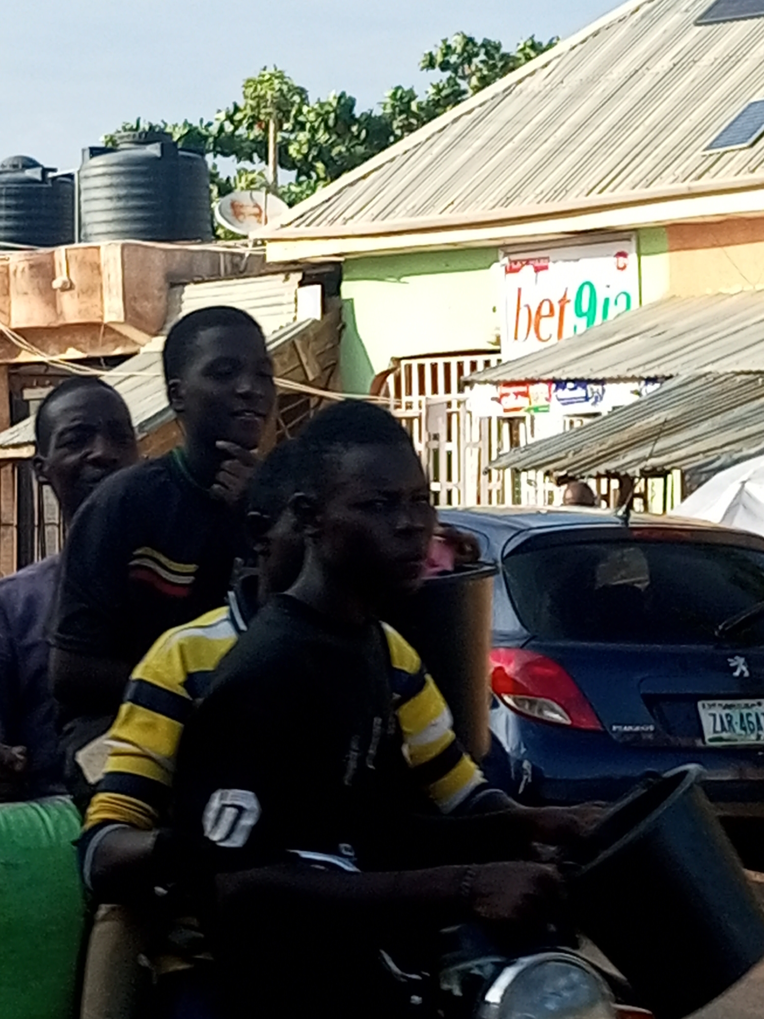 In Gombe, Nigeria, a group of young men are seen on a motorcycle. The rider is wearing a black and yellow striped shirt and holding a black bucket. Behind him, two other young men are seated, with one in the middle looking to the right and the other in the background looking forward. The scene takes place in an urban setting with a building visible in the background, featuring a corrugated metal roof and a sign that reads "bet9ja". Water tanks are also present on top of the building. A blue car with a license plate reading "ZAR 46A7" is parked behind the motorcycle. The lighting suggests it is daytime, possibly late afternoon.