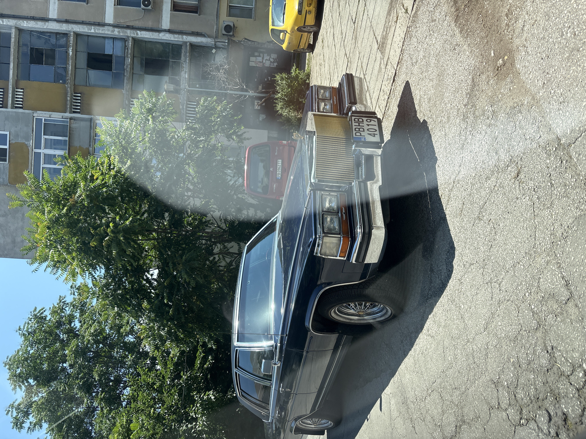The image shows a street scene in Plovdiv, Bulgaria, on a sunny day. In the foreground, a dark blue Cadillac sedan is parked on asphalt. The car has chrome accents and wire wheels. A Bulgarian license plate with the characters "PBHB 4019" is visible on the front. Behind the Cadillac, a red Citroen van is parked. Further back, a yellow car is visible. The background features a multi-story apartment building with balconies and windows, and lush green trees. The overall impression is of a residential area in an urban environment.
