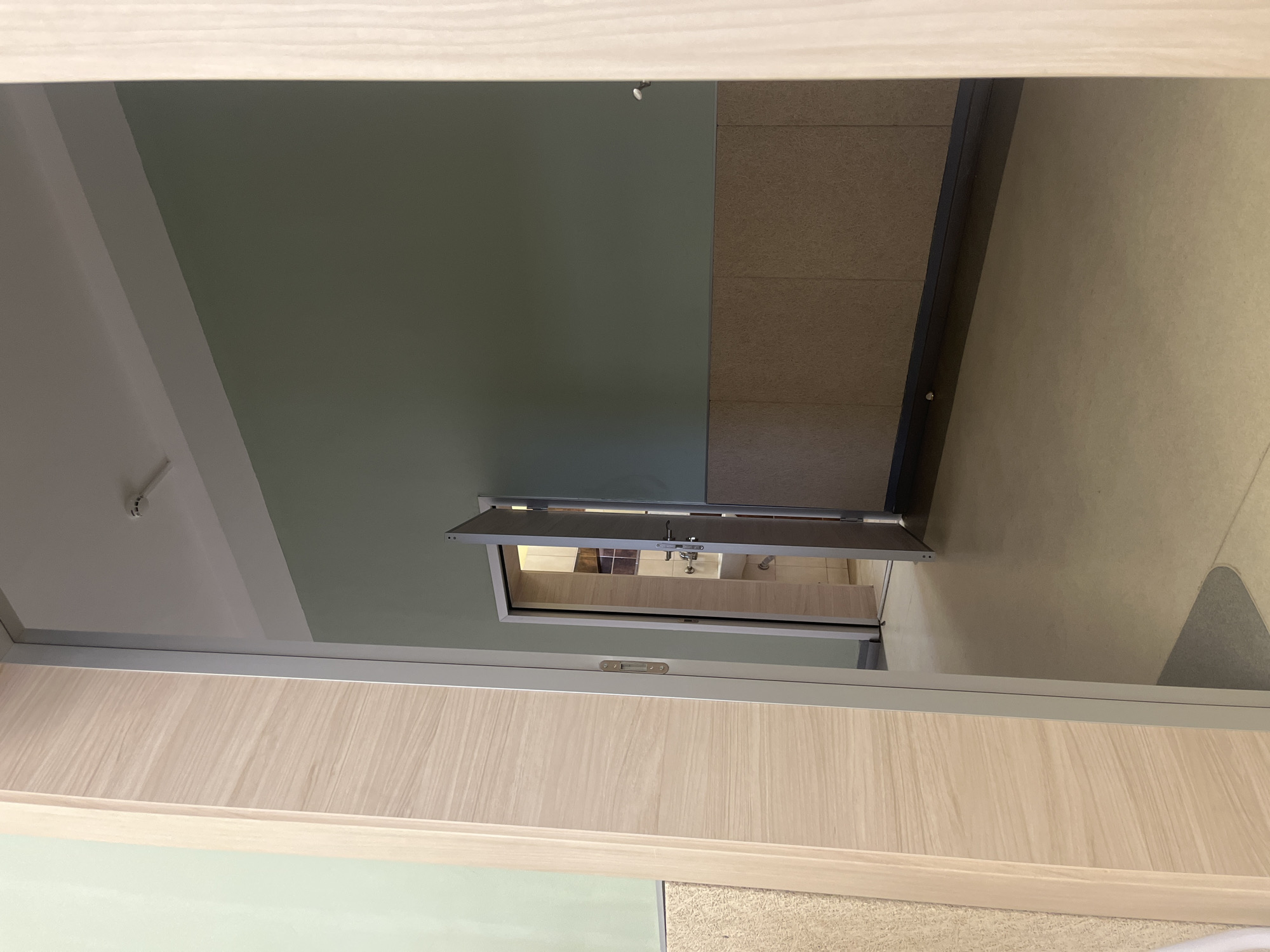 The image shows an interior view of a room, possibly in Plovdiv, Bulgaria. The perspective is from a lower angle, looking up towards the ceiling and a small open hatch or window. The ceiling is painted in two shades of green and white. The walls are covered with a textured, light brown material, resembling acoustic panels. Through the open hatch, there is a view into another room, possibly a bathroom, with beige tiles and plumbing fixtures visible. The frame of the hatch and the lower part of the image include wooden panels of a light brown color, possibly part of a cabinet or wall structure. No people are visible in the image. The overall environment appears to be an office or institutional setting.