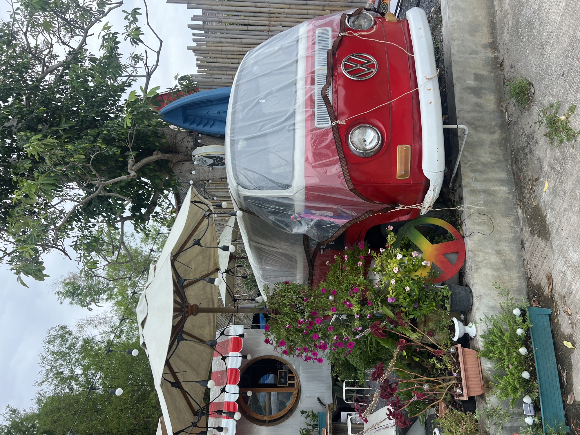 The image captures an outdoor scene featuring a red vintage Volkswagen Type 2 (VW Bus) serving as a prominent decorative and possibly functional element. The bus is positioned vertically in the frame, partially covered by clear plastic sheeting, and features a white bumper and the distinctive VW logo on its front. One of its visible wheels is creatively painted with a yellow, green, and red peace sign design.
To the left of the bus, a large, cream-colored patio umbrella with string lights attached provides shade. Beneath the umbrella is a small structure with a unique circular wooden-framed window. A sign inside this window clearly displays the text "Thai food," indicating the nature of the establishment.
The area around the bus and the structure is richly decorated with numerous potted plants and hanging baskets, overflowing with vibrant pink and purple flowers, contributing to a lush, garden-like atmosphere. Small white solar-powered garden lights are visible among the plants on the concrete ground, which also shows some cracks and weeds.
In the background, a blue kayak or small boat is turned upside down, resting on a surface behind the bus. A bamboo fence or wall provides a backdrop, and abundant green trees and foliage are visible throughout, suggesting a tropical or warm climate. The sky is overcast, indicating daytime with soft, diffused lighting. The scene conveys a quirky, inviting, and casual ambiance, likely a unique cafe or food stall.