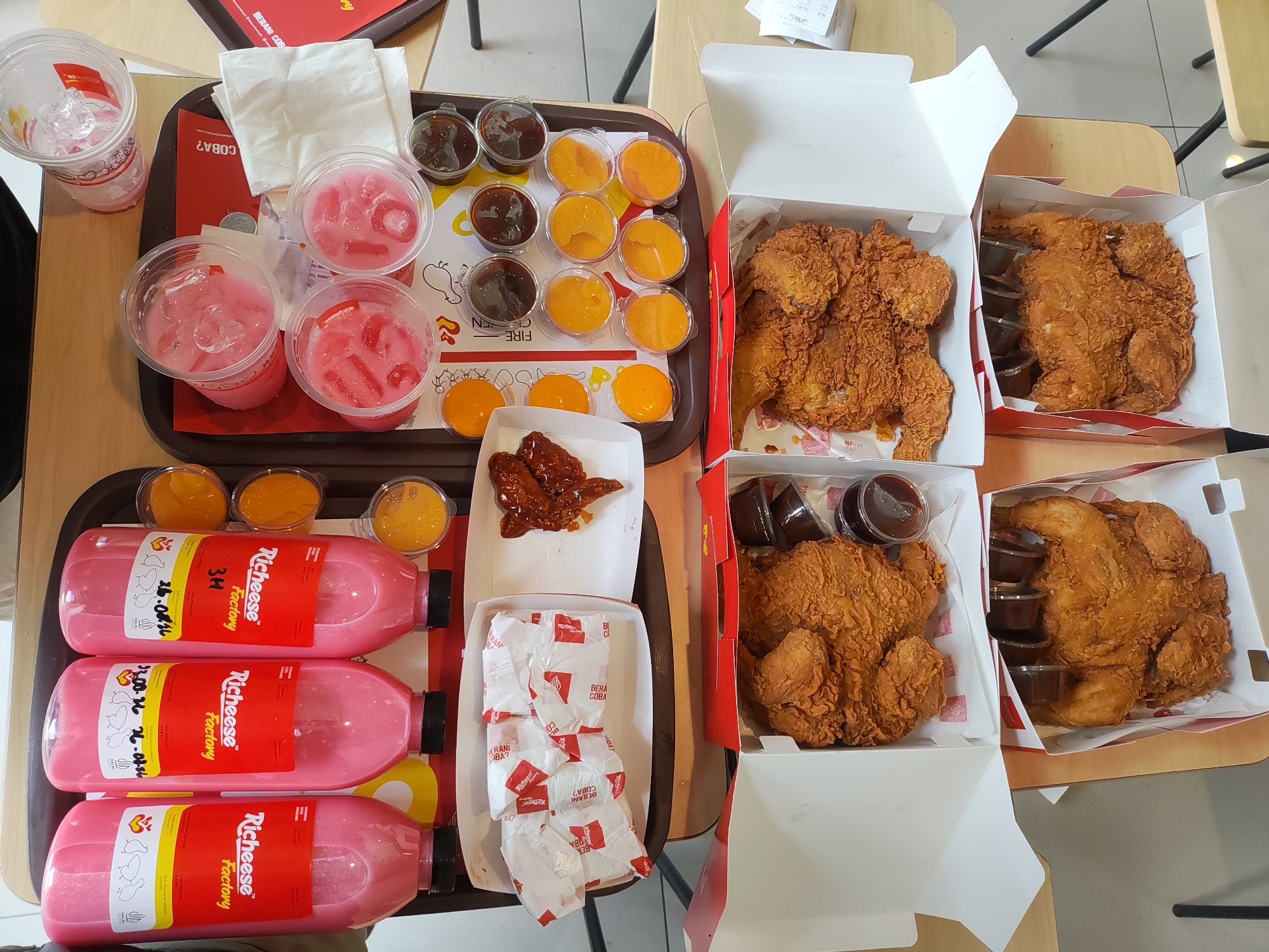 A tray of fast food items, including fried chicken, beverages, and dipping sauces, is displayed on a wooden table. Three bottles of pink beverage, labeled "Richeese Factory," are positioned vertically on the left side of the tray. Above these bottles are several cups containing pink drinks with ice, and a disposable cup with ice. To the right of the drinks, multiple small containers hold dark brown and orange dipping sauces. The tray also contains a small white box with fried chicken wings covered in sauce. Two larger white boxes, each filled with a whole fried chicken, are placed on the right side of the tray. The scene is set on a wooden table, with some menu items visible in the background. The overall arrangement suggests a mealtime setting, likely at a fast-food establishment.