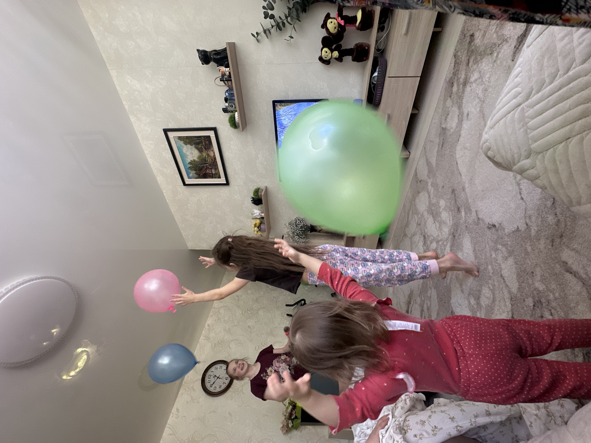 The image appears to be taken from a high angle, looking down into a room. It is a residential scene, likely inside an apartment or home.
There are three people visible: two children and an adult. One child is wearing red pajamas with white polka dots, and another is wearing a black top and patterned pants. The adult is wearing a maroon-colored shirt. The children are reaching for balloons, of which there is a large green one and a pink one. A blue balloon is visible near the ceiling.
The room contains a television set, a cabinet, decorative shelves with objects, a picture frame on the wall, and a clock on the wall. There is a ceiling light fixture.
There is not enough information to determine the exact location or determine if the picture was taken in the city of Uva, Russia.