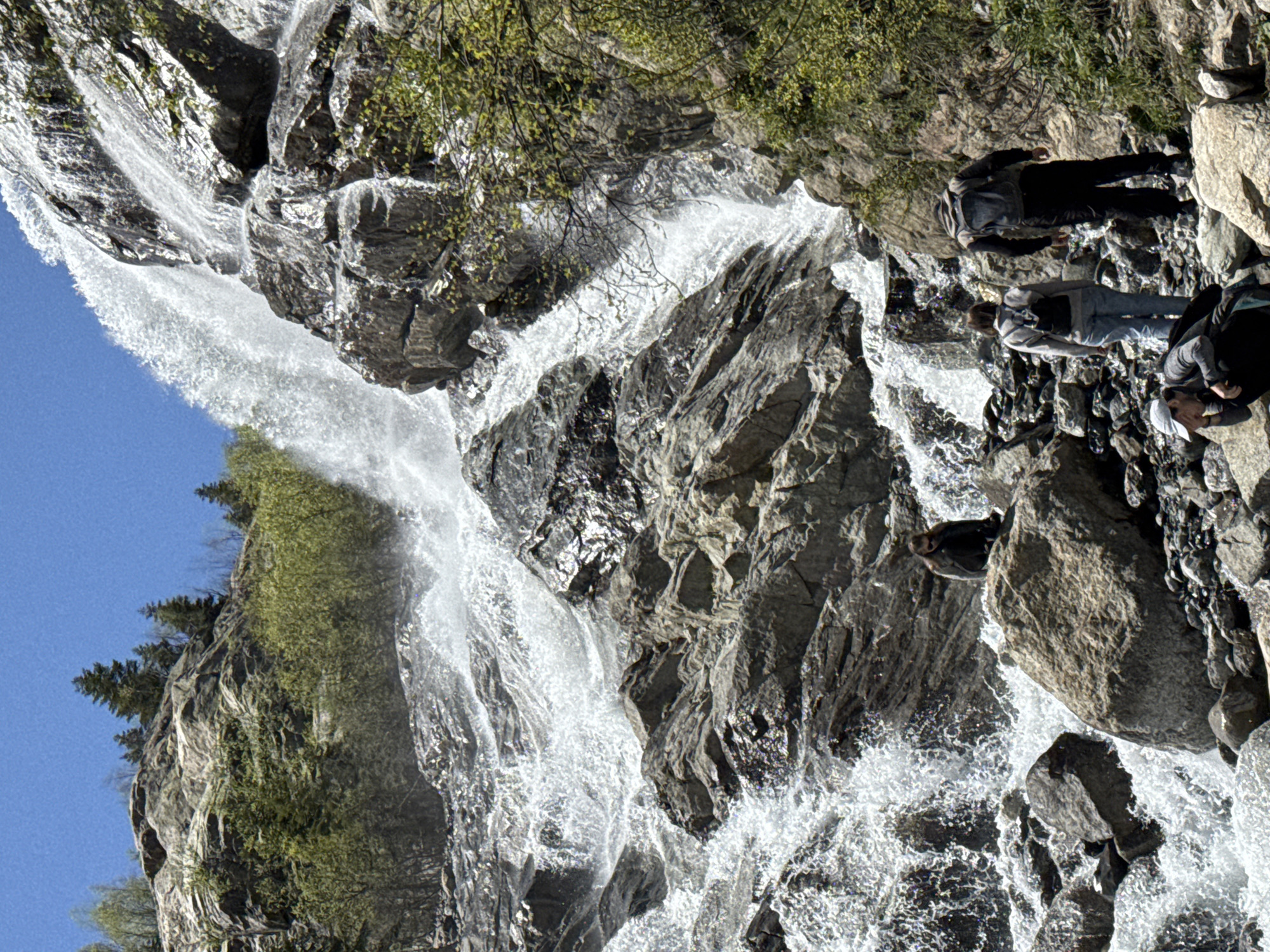 This full-frame image captures a multi-tiered waterfall cascading down a steep, rocky mountain slope under a clear blue sky in Верхне-Тебердинское сельское поселение, Russia. The waterfall dominates the center and right side of the frame, with white, frothy water turbulent over dark grey rocks and boulders. Sparse green vegetation, including coniferous and deciduous trees and bushes, is present on the rocky slopes surrounding the waterfall.
In the foreground, at the base of the waterfall, four individuals are distributed across a rocky terrain. On the bottom left, one person wearing a white baseball cap and dark clothing is crouched, facing downwards, with a dark backpack visible beside them. To the right of this individual, two people are walking upwards on a rocky path. The person in front wears light blue jeans, a light grey top, and a black backpack, while the person behind them wears dark pants, a dark top, and a grey backpack. Further left and slightly elevated, another person sits on a large grey boulder, facing left, wearing sunglasses and a dark jacket, observing the scene. The overall lighting suggests bright sunlight.