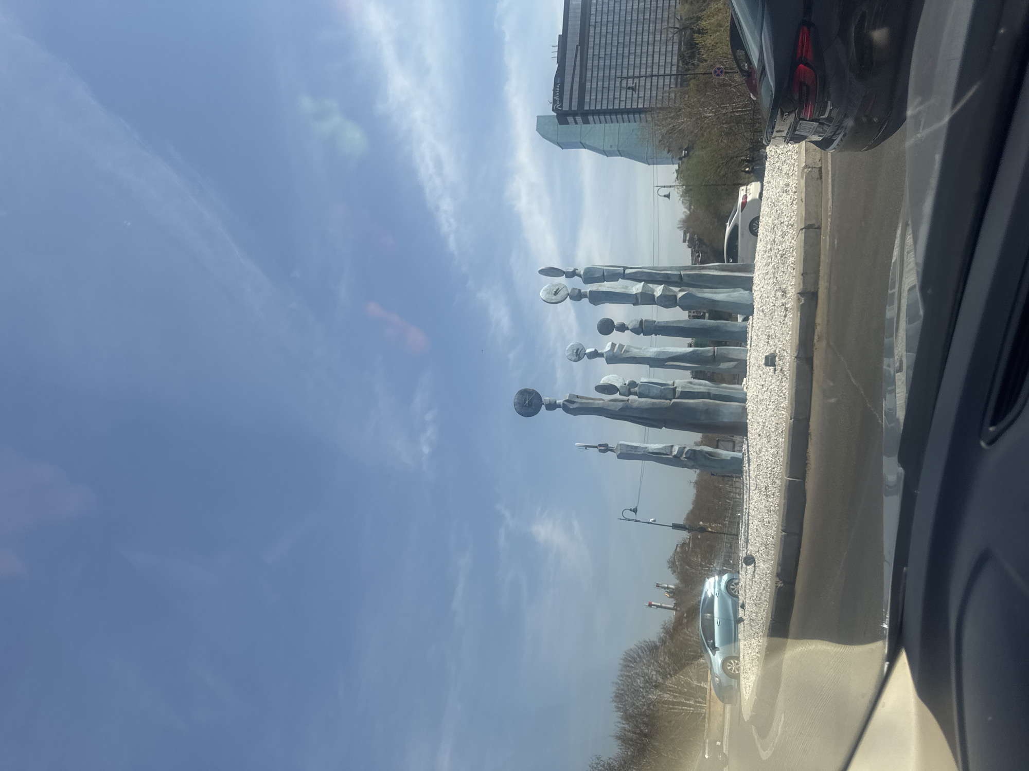 The image depicts a view from inside a car, likely a passenger seat. The focus is on a roadside sculpture in Tbilisi, Georgia, featuring a row of stylized figures with clock faces. To the right, a dark-colored car is partially visible. In the background, a tall building, possibly the Radisson Blu hotel, is visible against a blue sky with scattered clouds. The roadside features a curb made of light-colored stones. A light-blue car is seen further down the road. The overall scene suggests a sunny day in an urban setting.