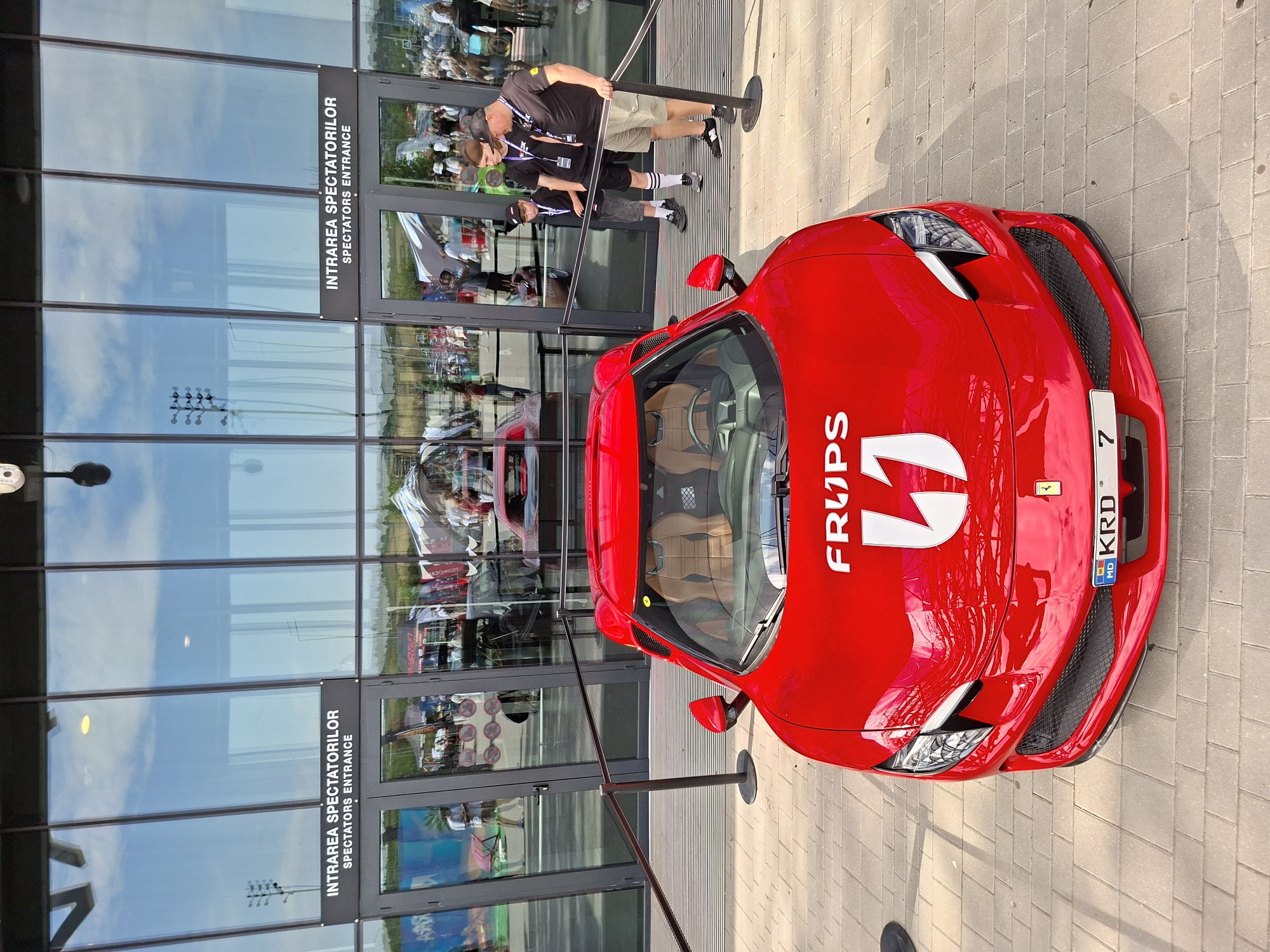 In Stăuceni, Moldova, a bright red sports car, possibly a Ferrari given the emblem on the license plate, is parked on a paved walkway in front of a modern building with large glass doors and windows. The car has tan leather interior and "FRUPS 1" is printed on the hood in white lettering. Above the car, a sign reads "INTRAREA SPECTATORILOR SPECTATORS ENTRANCE" in both Romanian and English, indicating the building is an entrance to a venue or event. Three individuals, two adults and a child, are standing to the right of the car, looking at it. The background shows a glimpse of an outdoor event with tents and other vehicles visible through the glass. The sky is visible through the glass, with clouds and what appear to be stadium lights in the reflection.