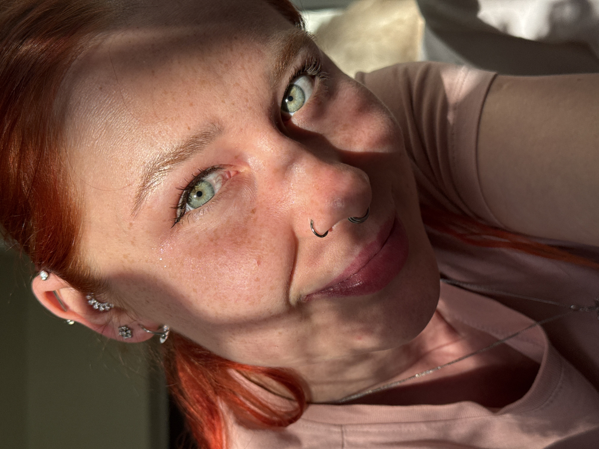 A close-up, upward-angled photograph shows a fair-skinned woman with red hair and green eyes. She has freckles on her face, a nose ring, and multiple piercings in her left ear, including a conch piercing, helix piercing, and lobe piercings. She is wearing a pink t-shirt and a silver necklace. Part of the scene is lit by bright sunlight. The location is not determinable from the image.