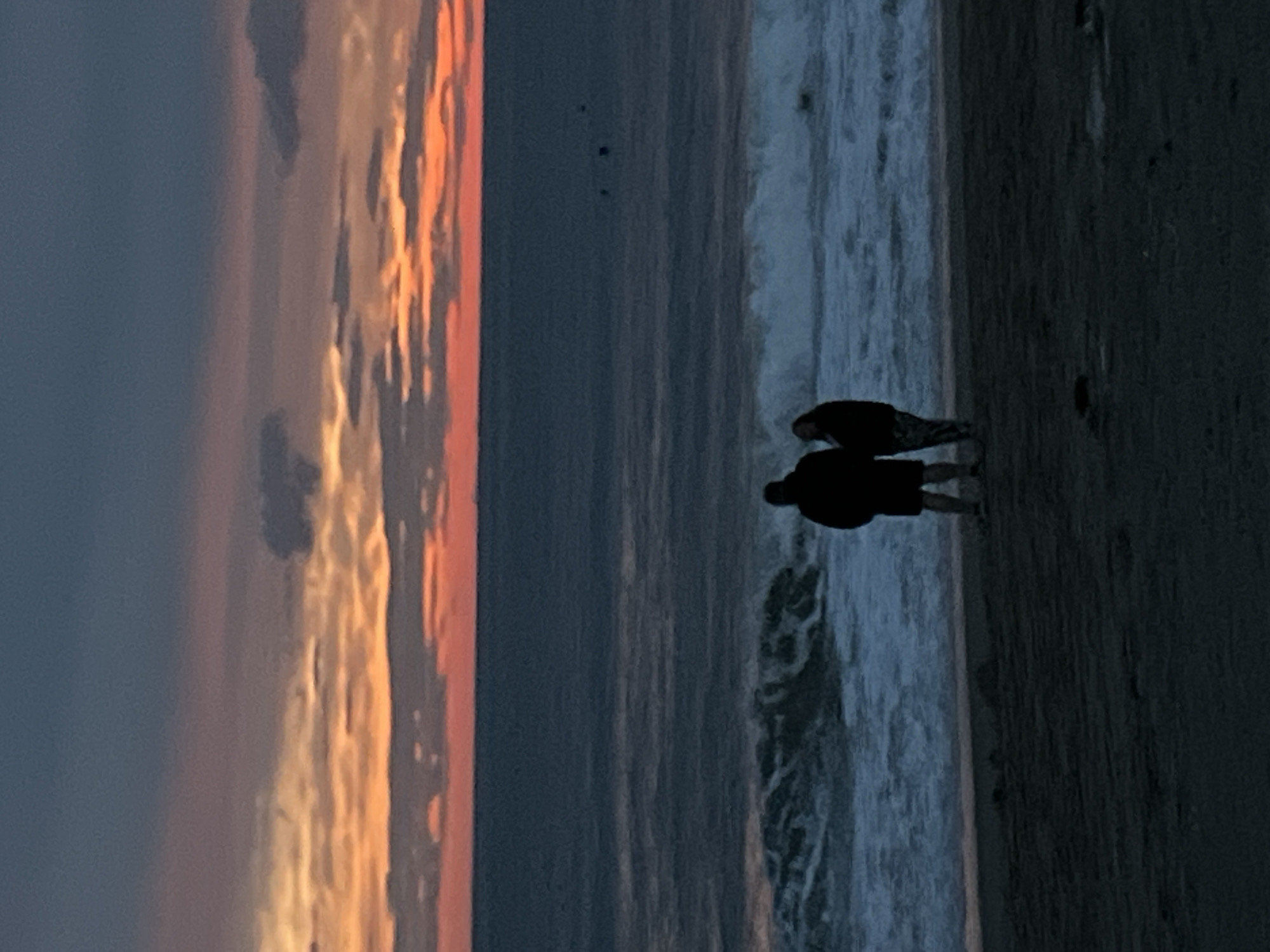 Here is a description of the image:
The image depicts a beach at sunset, likely in Alanya, Turkey. The sky exhibits a gradient of colors, from orange and pink near the horizon to darker shades of gray. A body of water extends to the right of the frame, displaying waves breaking on the shore. Two dark silhouettes stand on the sand. The sand itself is a dark color and has a grainy texture.