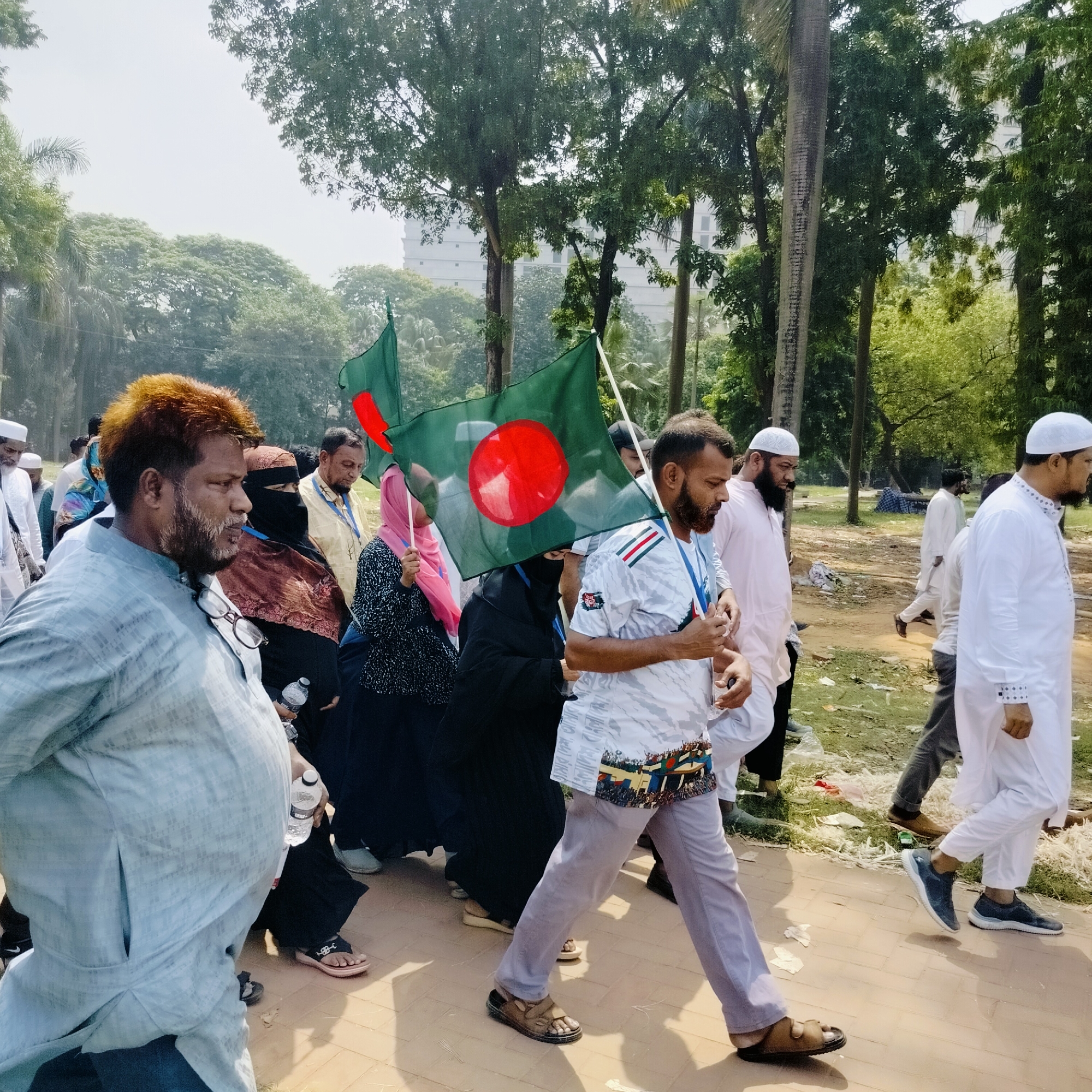 A group of people are walking along a paved path in what appears to be a park or public space. The group includes both men and women, some of whom are wearing traditional Muslim clothing such as hijabs and skullcaps. Several individuals are carrying the flag of Bangladesh. The environment consists of trees, green foliage, and a building in the background, consistent with an urban park setting. The location is likely Dhaka, Bangladesh, based on the presence of the national flag and the style of clothing.