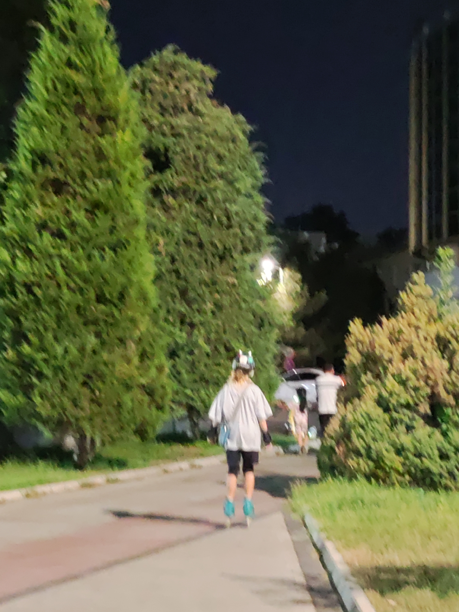 The image captures a nighttime scene on a paved pathway lined with lush green trees and grass. In the foreground, a person is rollerblading away from the camera. They are wearing a light-colored hoodie, dark shorts, and blue rollerblades. A light blue bag is slung across their shoulder. Behind the rollerblader, further down the path, two other individuals are visible, one in a white shirt and another in a patterned dress, with a white car parked nearby. To the right of the path, a dense, yellowish-green shrubbery stands out, and to the left, tall, dark green evergreen trees dominate the landscape. In the background, a tall, multi-story building with vertical lines is partially visible, illuminated by the surrounding lights. The overall ambiance suggests a recreational area within a city, possibly a park or a well-maintained urban pathway in Tashkent, Uzbekistan, given the context.