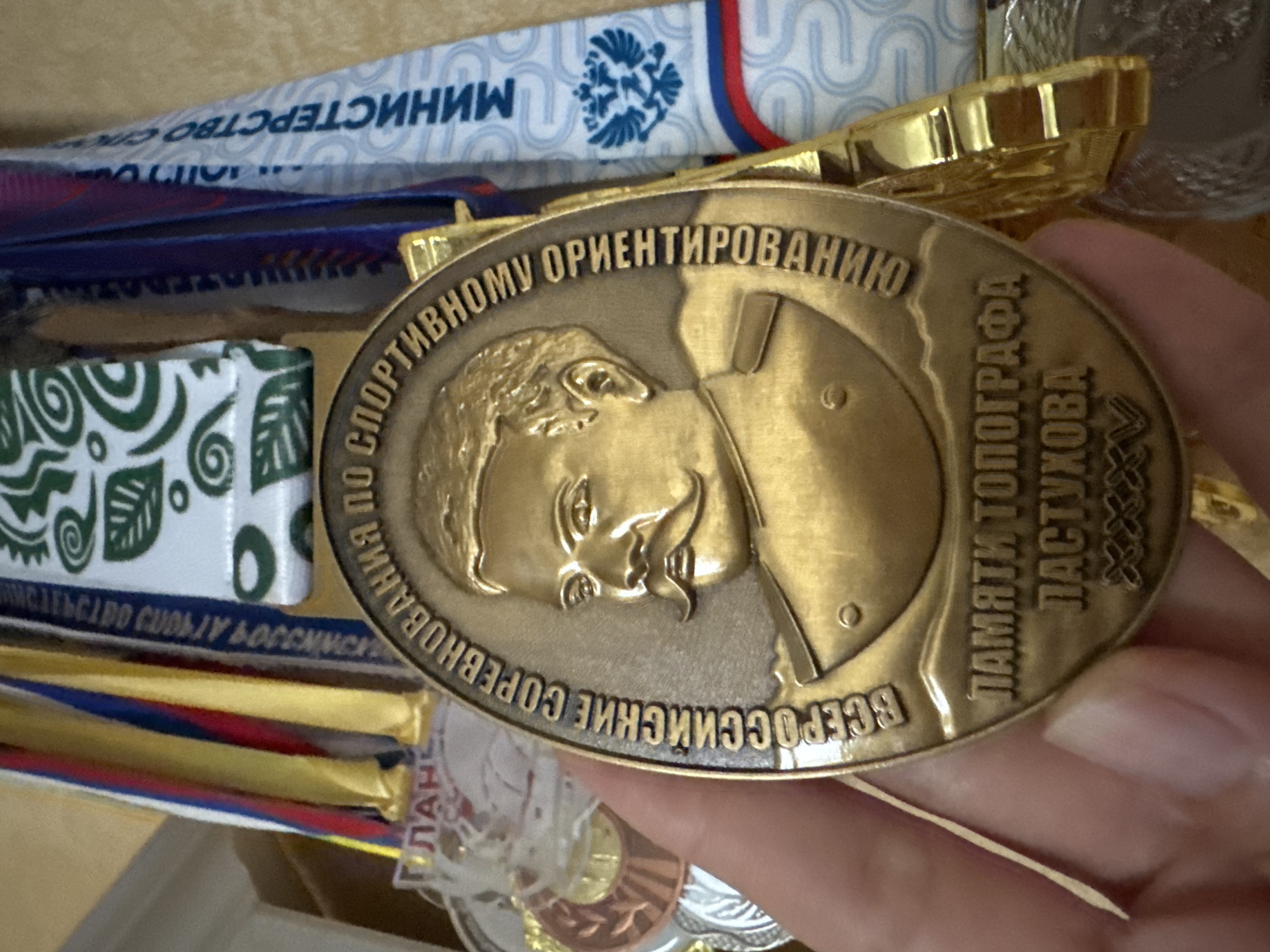 The image shows a close-up of a hand holding a gold medal. The medal features a relief portrait of a man with a mustache and epaulets, surrounded by text in Russian. Below the portrait, more Russian text is visible, along with what appears to be Roman numerals. The medal is suspended by ribbons of red, blue, and yellow. In the background, other medals and ribbons are partially visible, including one with a blue and white pattern and a Russian double-headed eagle emblem. The lighting suggests an indoor setting. The medal text translates to: "All-Russian Competitions for Topographer Pastukhov" and then Roman numerals "XV". There is no visible indication of the location being the city of Sinyavino, Russia.