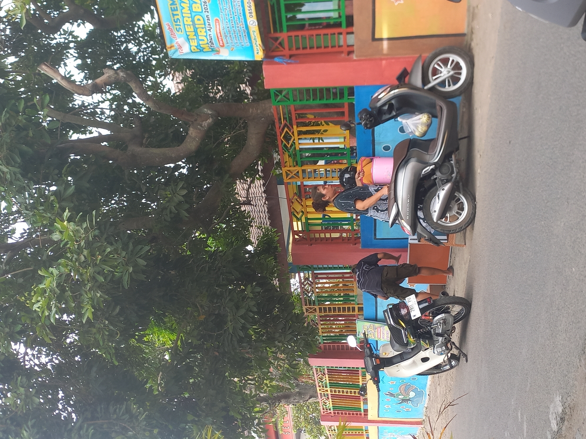 The image depicts a street scene in front of a building with a colorful fence. Two motorbikes are parked on the side of the road. A woman, wearing a patterned top and dark pants, stands next to a grey scooter, holding a pink bucket. Another person, wearing a dark shirt and khaki shorts, is bending down near a white motorbike. The building behind the fence appears to be a school, with a sign that reads "TELAH DIBUKA SISTEM PENERIMAAN MURID BARU TAHUN AJARAN 2026-2027". The area seems to be Tambak Rejo, Indonesia, given the contextual information provided. The environment is characterized by a large tree providing shade, a brightly painted fence, and a paved road.