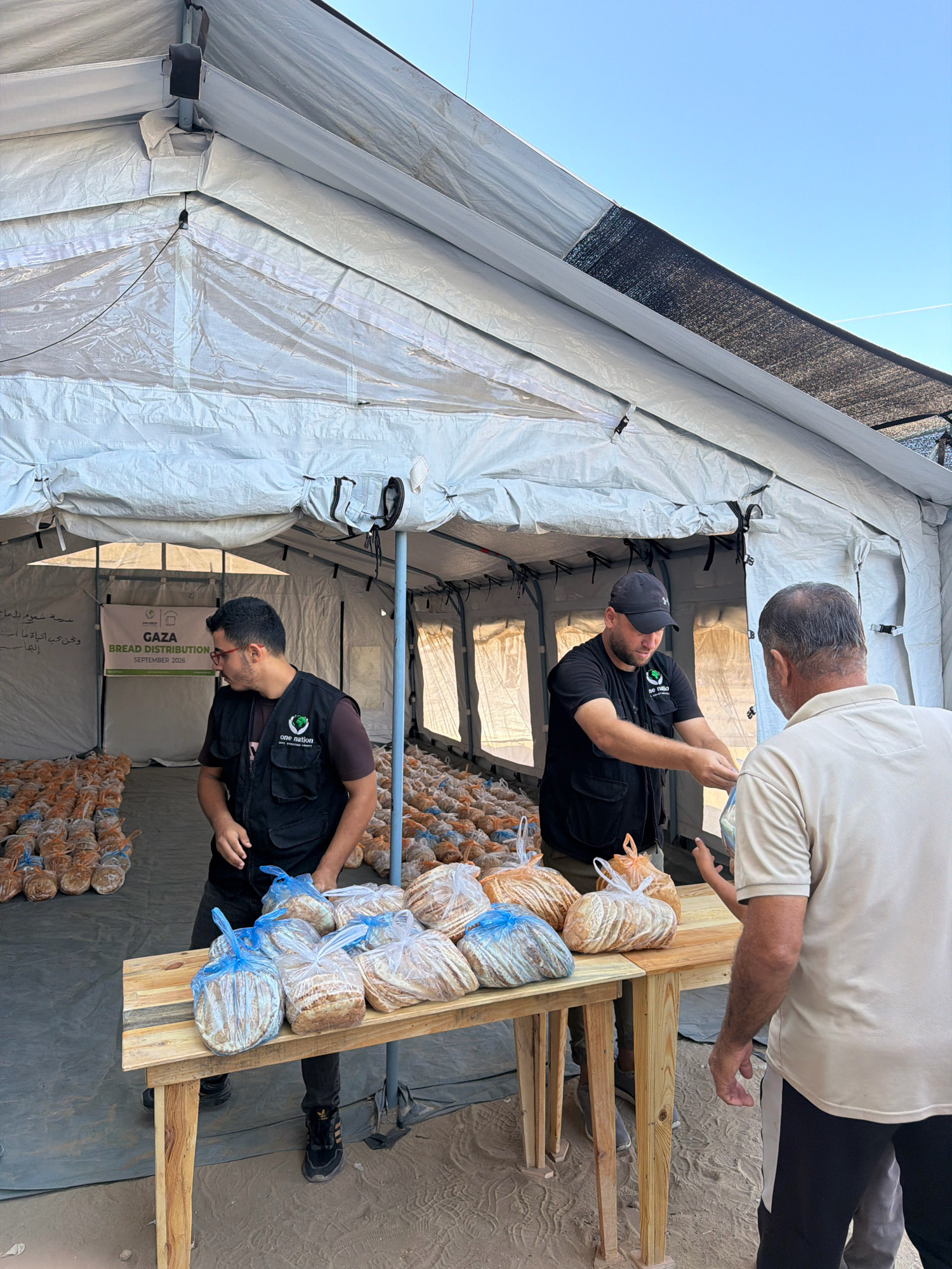 In the city of Gaza, Palestinian Territories, two individuals wearing vests with the logo "one nation" are distributing bread under a large white tent. One person, wearing a black baseball cap and a black vest over a black t-shirt, is seen handing a bag of bread to an older man wearing a light-colored polo shirt. Bags of bread are stacked in rows on the ground behind them, and more bread is placed on a wooden table in front of them. A banner in the background reads "GAZA BREAD DISTRIBUTION SEPTEMBER 2026." The ground appears to be sandy. The sky is clear and blue.