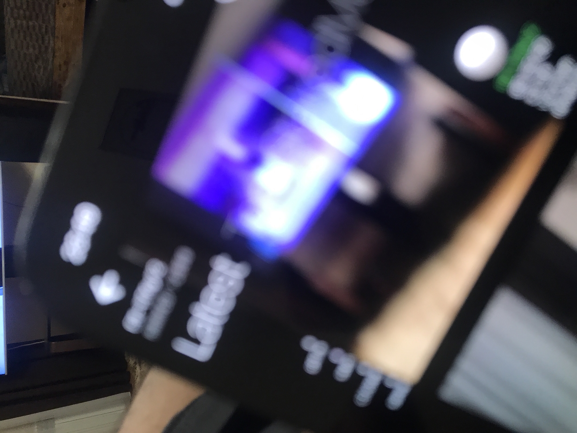The image shows a close-up of a smartphone screen held by a person. The screen displays a blurry image with a purple and white light source. Text and symbols are visible on the screen, although illegible due to the focus. The person's arm and possibly shoulder are visible, indicating they are holding the phone up. Elements in the background include a wooden shelf and a partially visible TV screen. The location in Yerevan, Armenia, is not discernible from the visual information available in the image.