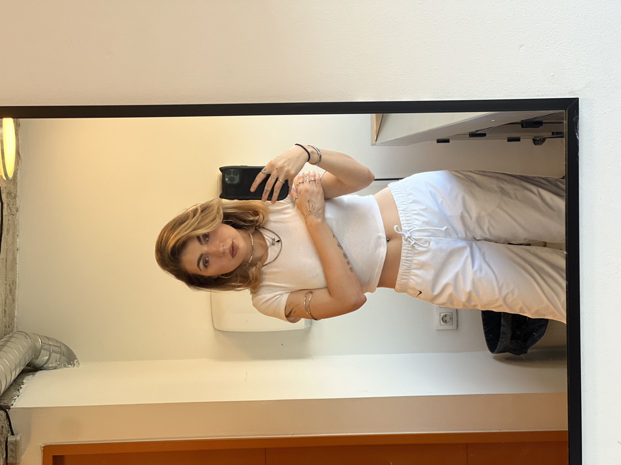 A young woman with blonde hair is taking a mirror selfie in what appears to be a public restroom. She is wearing a white crop top and white track pants. Her left arm is wrapped around her waist, and her right hand is holding her phone. The background shows a white wall, an orange door, a trash can, and electrical outlets. The mirror reflects the woman and the surrounding environment. The scene is not in Sofia, Bulgaria.