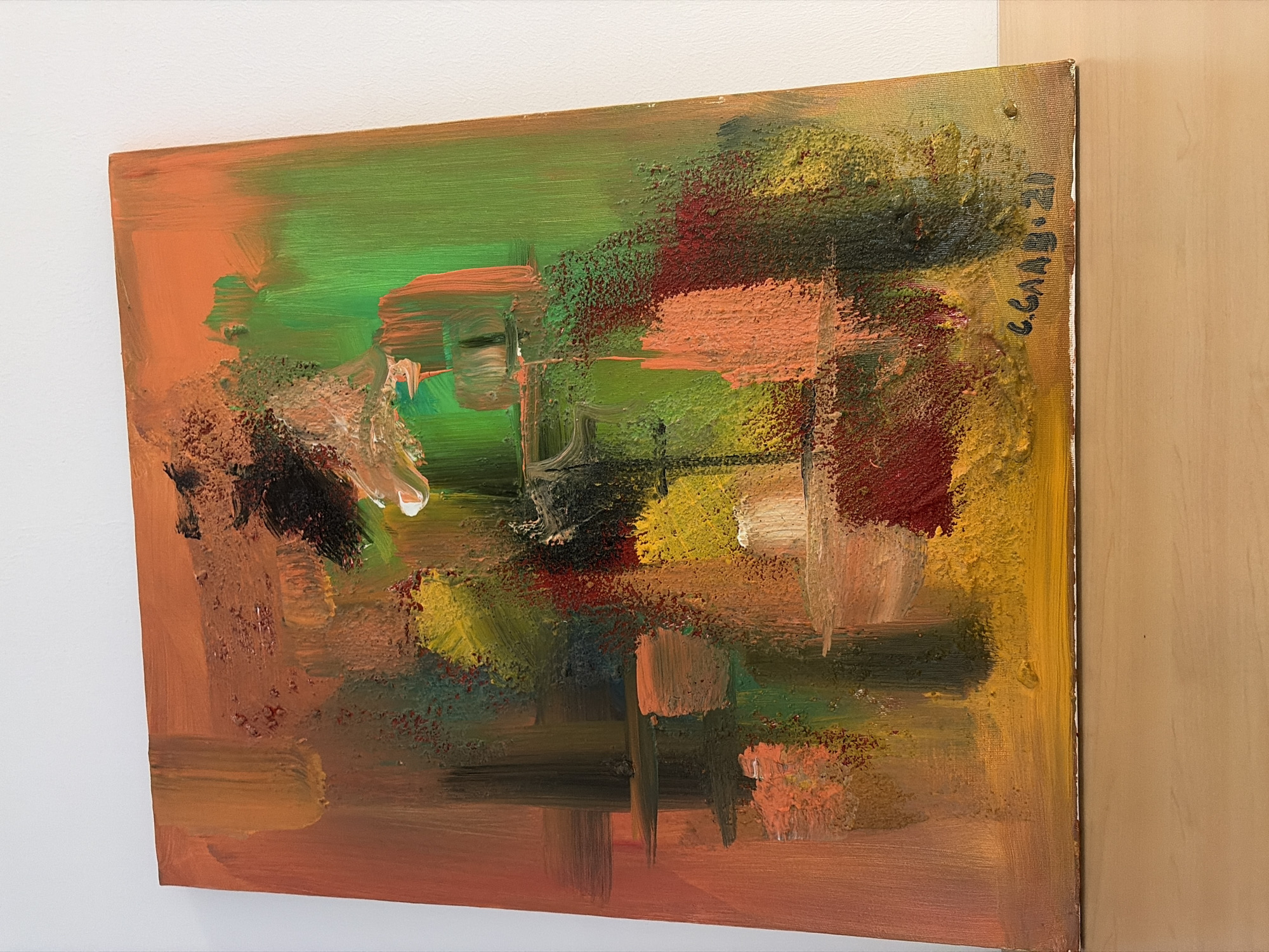 The image depicts an abstract oil painting hanging on a white wall. The painting features a blend of colors, including orange, green, yellow, red, and black. The painting also has a signature and "20" written on the right side. The painting is displayed indoors, adjacent to a wooden panel. There is no indication of Sofia, Bulgaria.
