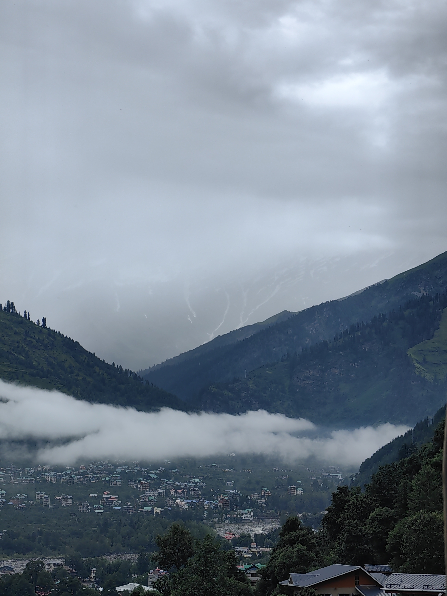 The image depicts a hazy, mountainous landscape with a village nestled in the valley. The sky is overcast with thick, grey clouds, and mist obscures the upper reaches of the mountains. In the foreground, lush green hillsides are dotted with trees. A band of white clouds hangs low in the valley, partially obscuring a cluster of buildings that make up the town of Vashisht, India. The buildings appear to be a mix of houses and hotels, with colorful roofs visible. The overall atmosphere is serene and slightly melancholic due to the cloudy weather. There are no visible people or animals in the image.