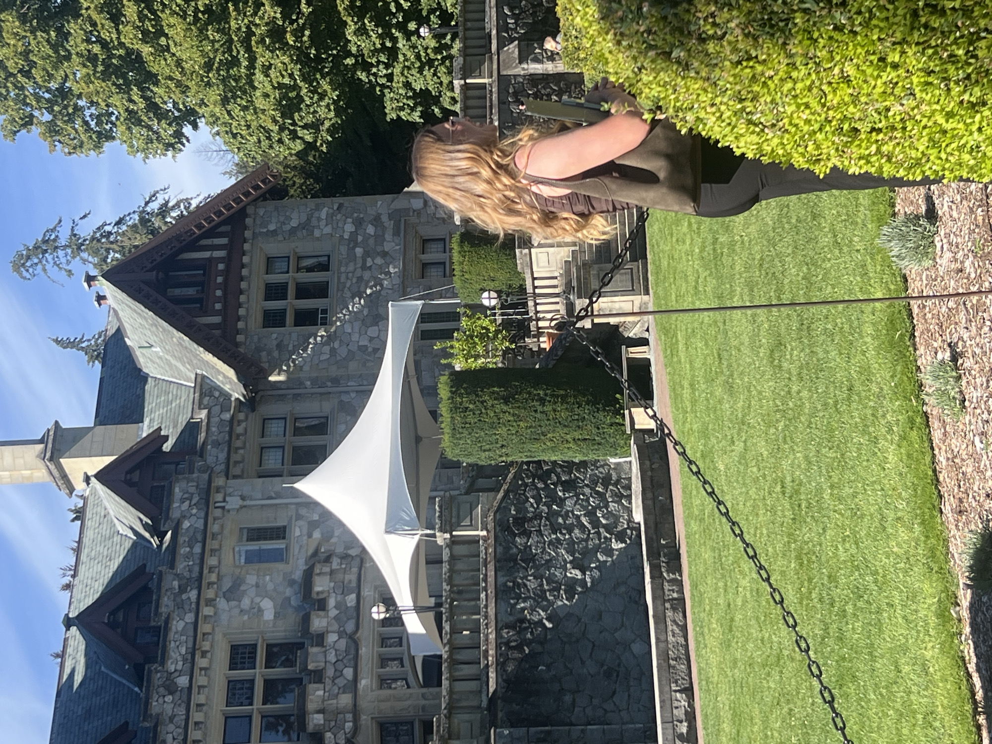 In the city of Langford, Canada, a woman with long blonde hair and sunglasses stands in front of a stone mansion. She is wearing a brown top and black pants, and is holding a green bag and a document. A white tent is set up on the lawn in front of the mansion. The mansion is a large, stone building with a dark roof and several windows. The lawn is green and well-manicured, with a chain link fence running across it. Tall trees are visible in the background. The sky is blue and clear. The scene appears to be a sunny day.