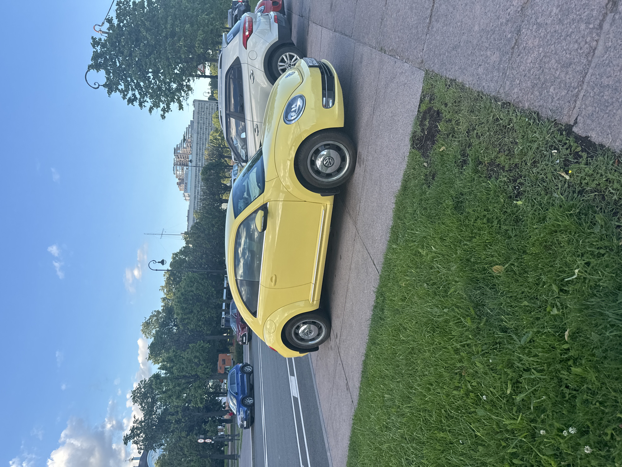 In Saint Petersburg, Russia, a yellow Volkswagen Beetle is parked on a paved sidewalk next to a grassy area. The car is positioned on the right side of the frame, with its front facing towards the right. In the background, other vehicles, including a blue SUV and a white crossover SUV, are visible on a road. A line of trees runs along the edge of the road, and beyond that, a multi-story building complex is visible under a clear blue sky with scattered clouds. A lamppost with a distinctive curved arm stands near the trees. The overall scene depicts a typical urban streetscape with vehicles and greenery.