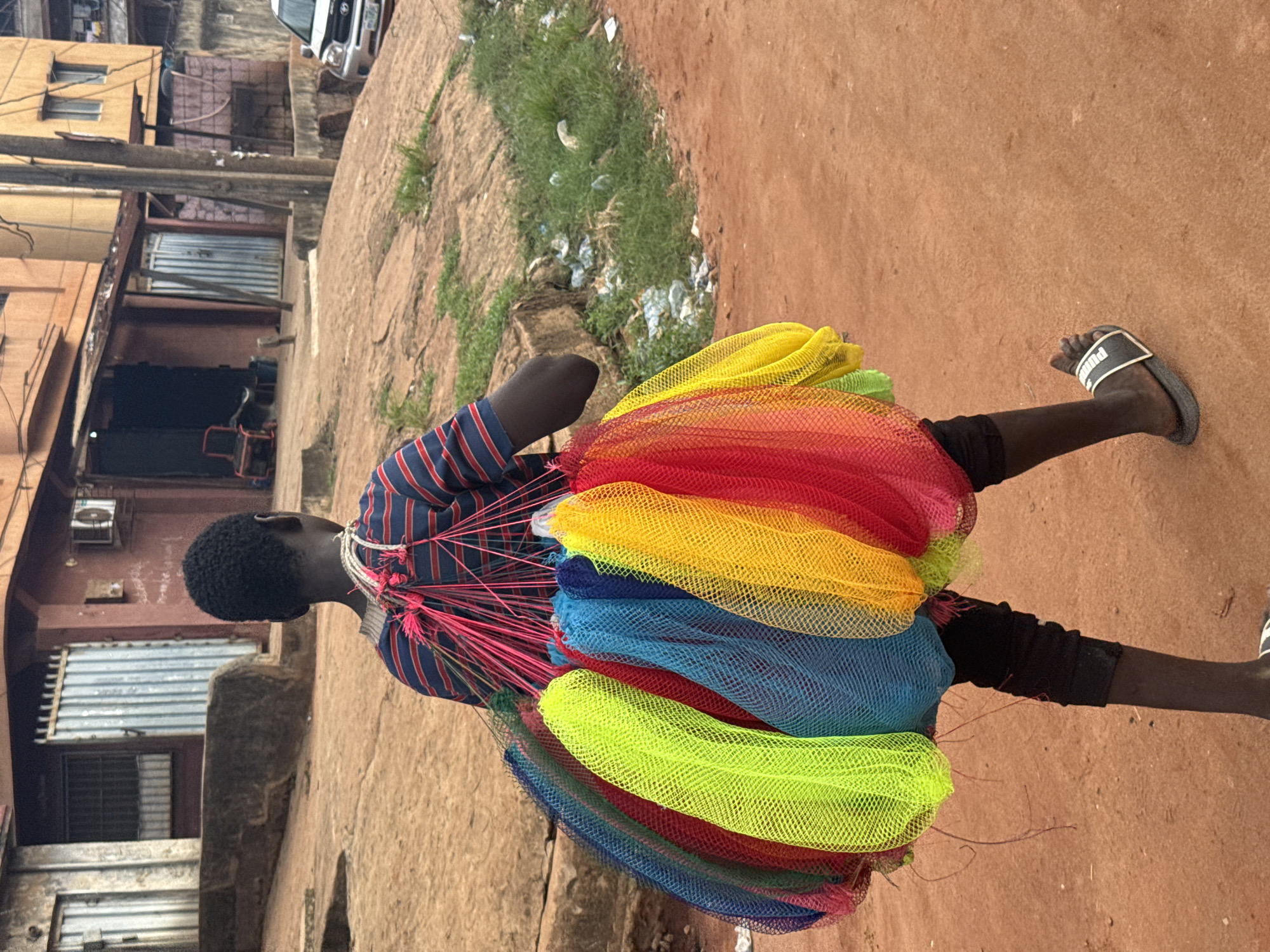 A young man stands on a red dirt road in Onitsha, Nigeria. He carries a large, colorful bundle of netting, in the colors of red, yellow, blue, and green. He wears a blue and red striped shirt, black shorts, and black and white sandals. The background shows a weathered building with metal shutters, a concrete pole, and a patch of grassy weeds. A vehicle is visible in the background.