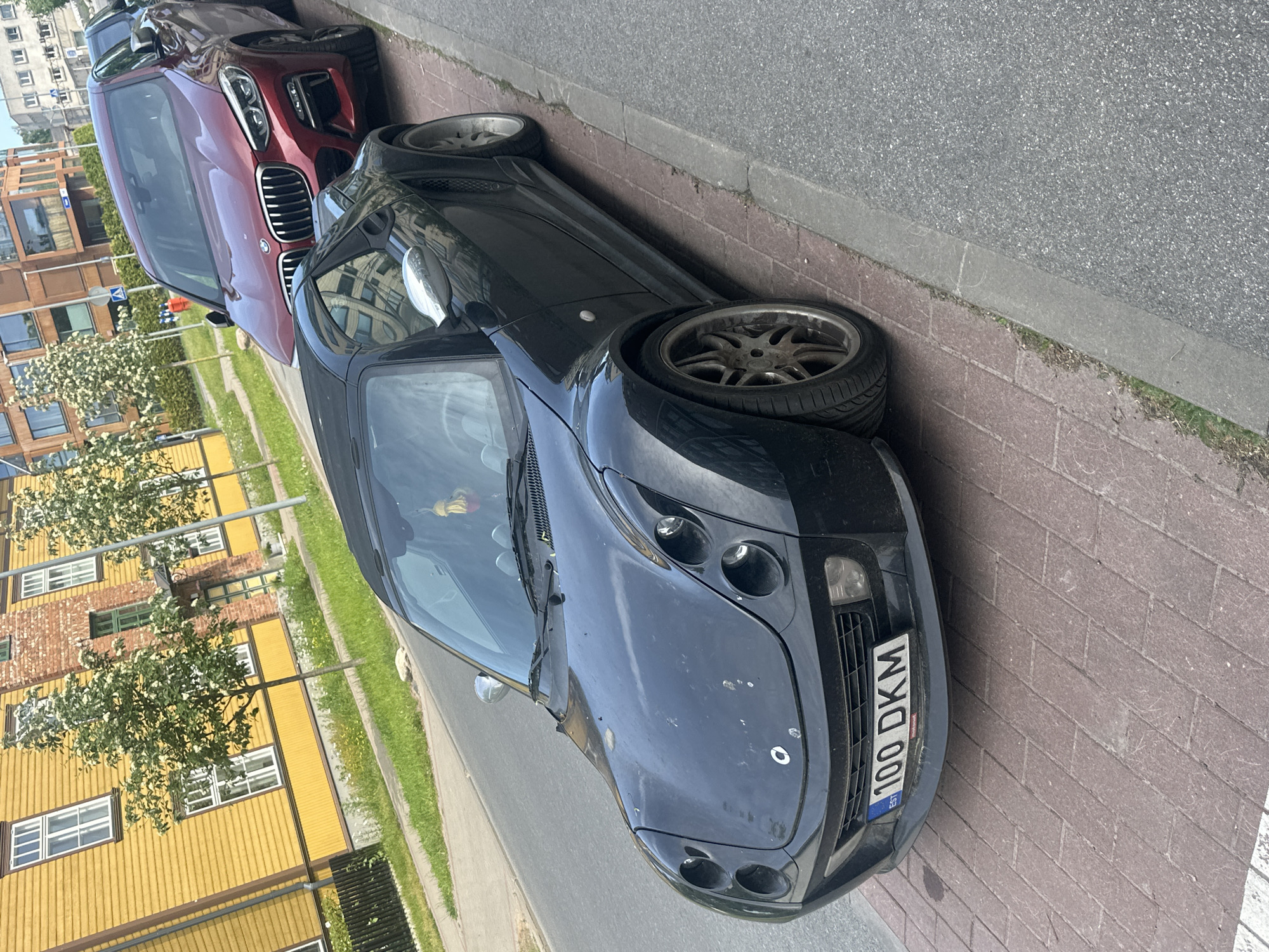 The image captures a street scene in Tallinn, Estonia, focusing on two parked cars. A black Smart Roadster is prominently featured in the foreground, its front bumper displaying an Estonian license plate with the characters "100 DKM". The car has a convertible roof. Behind it, a darker red BMW SUV is parked. Both vehicles are situated on a paved area next to a street. In the background, a yellow wooden building with white-trimmed windows and a brick section with green window frames suggests an urban residential or commercial area. Trees with white blossoms line the street, and modern apartment buildings are visible further in the distance. The ground is a mix of red brick paving and asphalt.