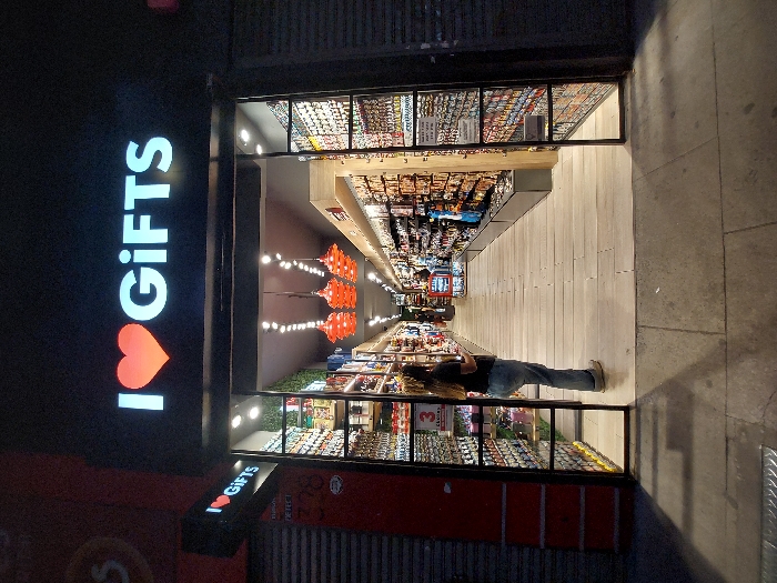 A vertically oriented image showcases the exterior of a brightly lit "I ❤️ GIFTS" store. The store is full of merchandise displayed on shelves along a long aisle, with red lanterns hanging from the ceiling. A person is browsing in the store. The shop has a black metal frame and is located on a street with a concrete sidewalk.