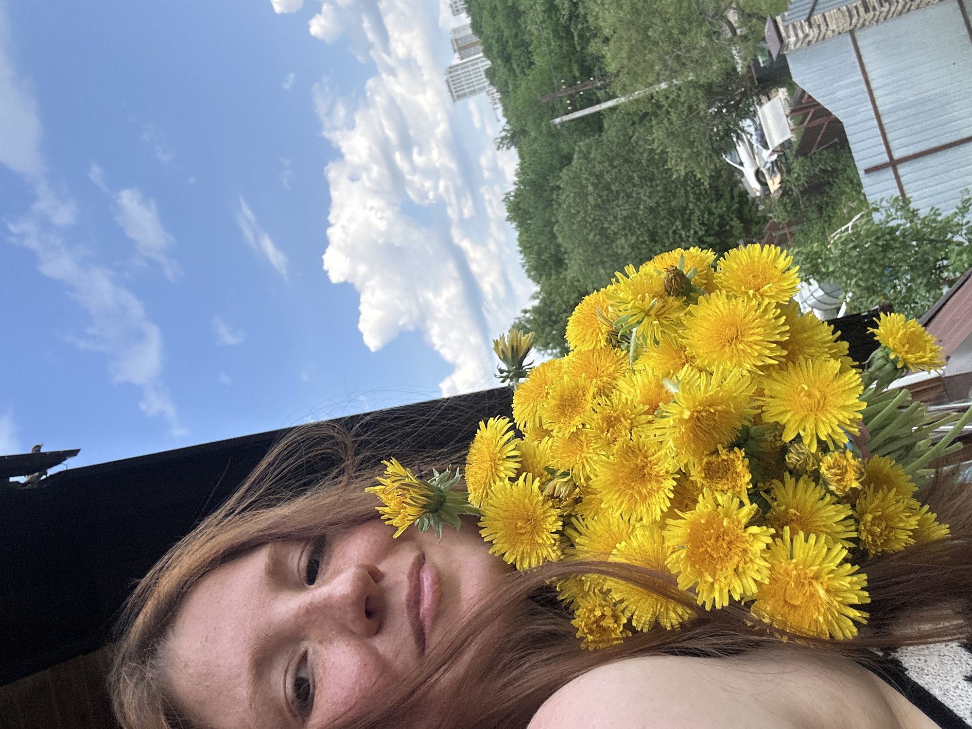 The image shows a woman with light brown hair holding a large bouquet of yellow dandelions. One dandelion is tucked into her hair near her forehead. Her face is partially visible, showing freckles and a neutral expression. The background features a blue sky with white clouds, as well as a line of trees and a white building in the distance. Part of a roof or structure is visible in the top right corner. Based on the building style and landscape visible, the photo likely originates from Nizhny Novgorod, Russia.