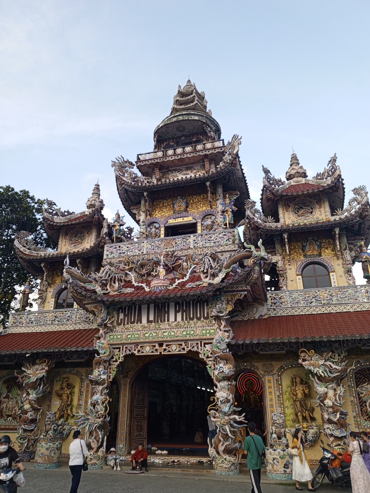 The image captures the exterior of Chua Linh Phuoc, a pagoda located in the city of Xuan Thanh, Vietnam. The pagoda is adorned with intricate, colorful mosaic tiles featuring dragons, Buddhist figures, and floral patterns. The architecture is characterized by multiple tiered roofs and ornate decorations. In the foreground, several people are present: a person on a scooter is entering from the left, a woman in a white shirt and dark pants stands near the entrance, and two people are sitting on the ground to the right of the entrance. Further inside the pagoda's entrance, more people are visible. Various objects like shoes and a motorcycle are also present. The sky above is clear and pale.