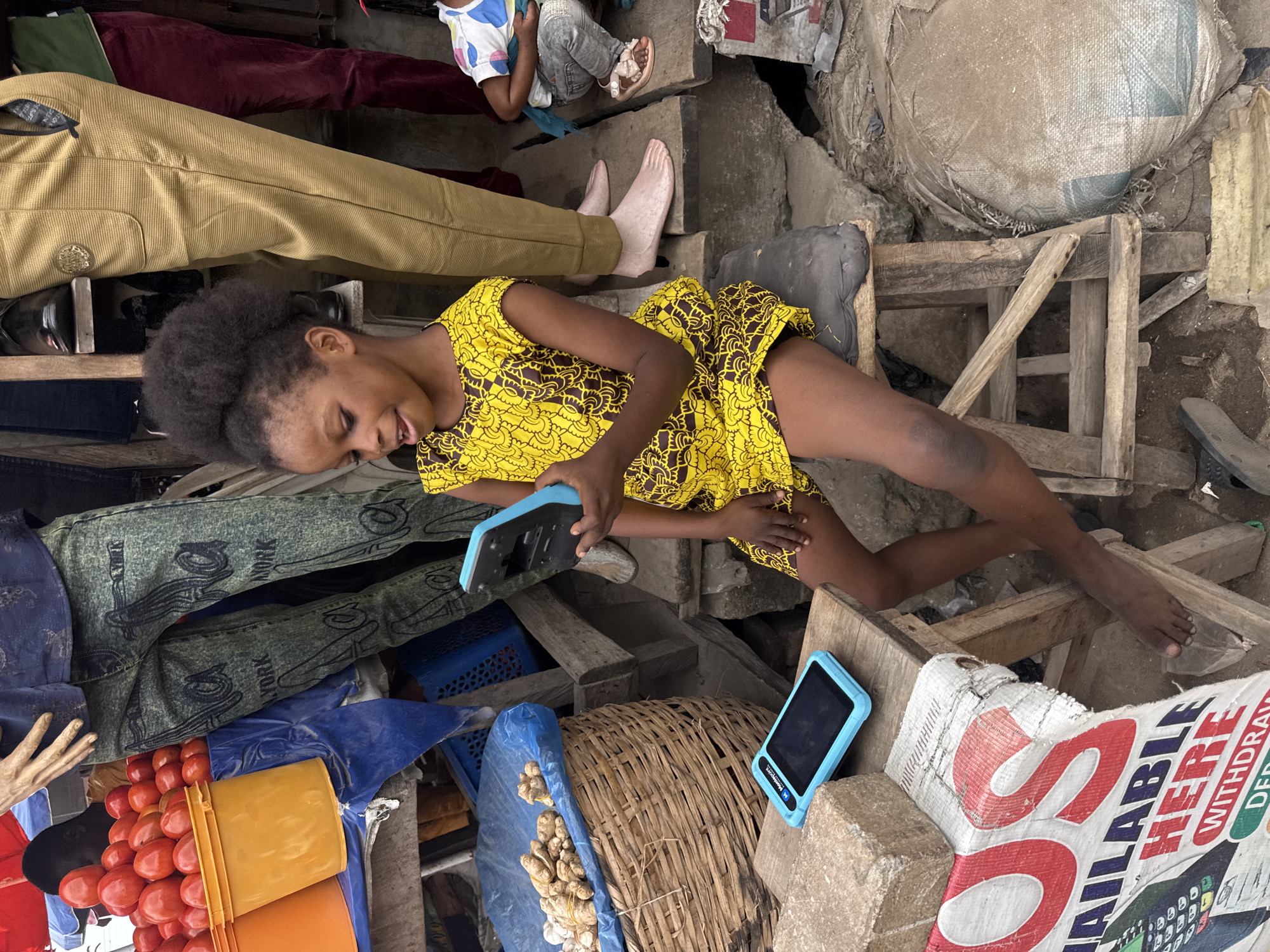 In Ado, Nigeria, a young Black girl, appearing to be around 6-8 years old, sits on a low wooden stool in a bustling market. She wears a vibrant yellow dress with a brown pattern and holds a blue mobile payment device in her hands, seemingly engrossed in it. To her left, on a makeshift wooden table, another identical device rests on a stone. The background reveals a chaotic yet active marketplace scene: stacked orange buckets filled with red tomatoes, a basket containing ginger, blue plastic crates, and sacks of goods. Several adults are partially visible, including one person in patterned green trousers and another in tan trousers. The ground is rough and unpaved. The overall impression is of a lively, everyday market environment in Nigeria.