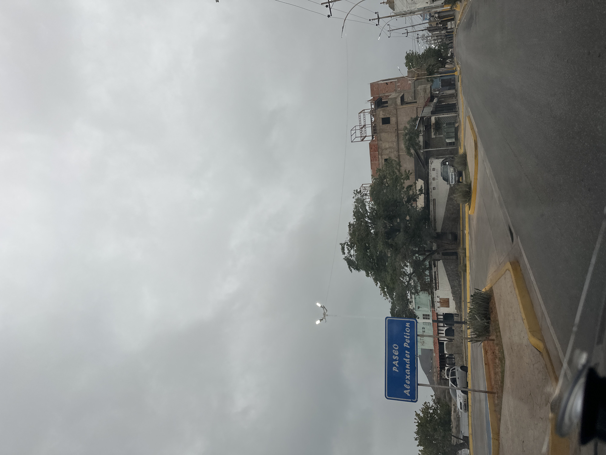 The image shows a street scene in Juan Griego, Venezuela, under an overcast sky. A blue street sign in the foreground reads "PASEO Alexander Petion". To the left of the sign, a street lamp with two lights is visible. Behind the sign, there are buildings, including one that appears to be under construction with exposed brick and a metal frame on the roof. A large tree with dense foliage is situated between the sign and the construction site. A silver car is parked on the left side of the street, and a dark SUV is parked further down. The street is paved with asphalt and has yellow curbs. In the background, more buildings and utility poles line the street. The overall atmosphere is muted due to the cloudy weather.