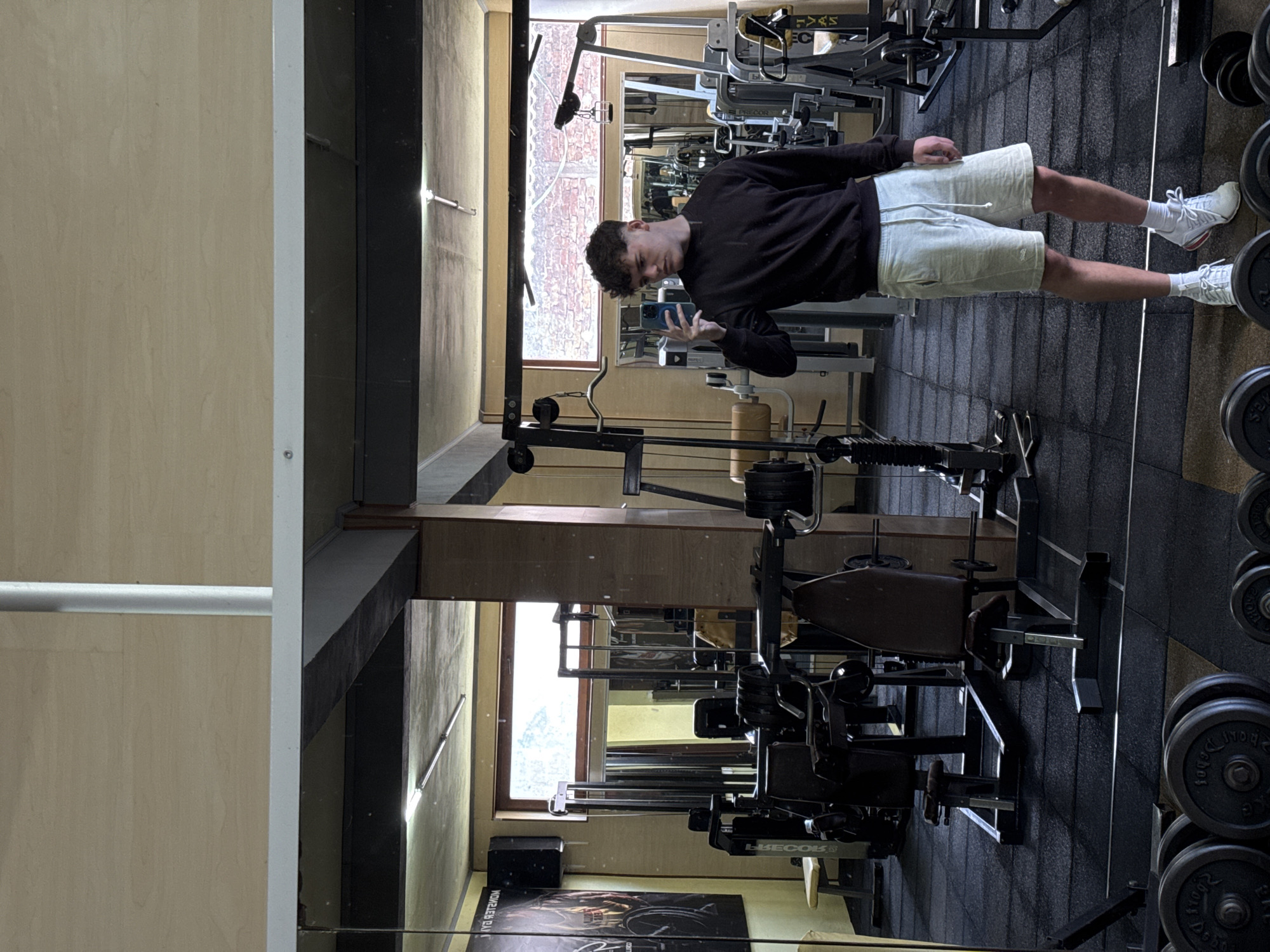A young man is standing in a gym in Goce Delcev, Bulgaria, reflected in a mirror. He is wearing a black long-sleeved shirt, light-colored shorts, and white sneakers. He is holding a smartphone and appears to be taking a picture of himself in the mirror. The gym has various weight machines and dumbbells. There is a window reflecting a building across the street. The walls of the gym are painted a light yellow, and the floor is covered in black rubber tiles.