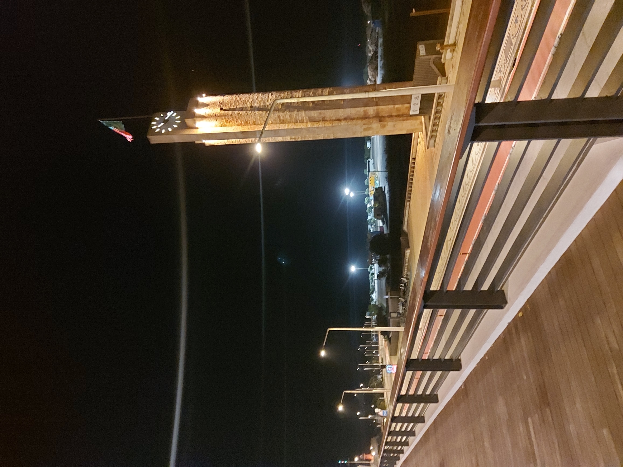 The image shows the night view of the Ponte Edgar Cardoso bridge in Figueira da Foz, Portugal. The bridge is illuminated, and its structure is visible, including the beams and supports. A clock and a flag are seen on the bridge's side. Streetlights are also visible along the roadway. The sky is dark.
