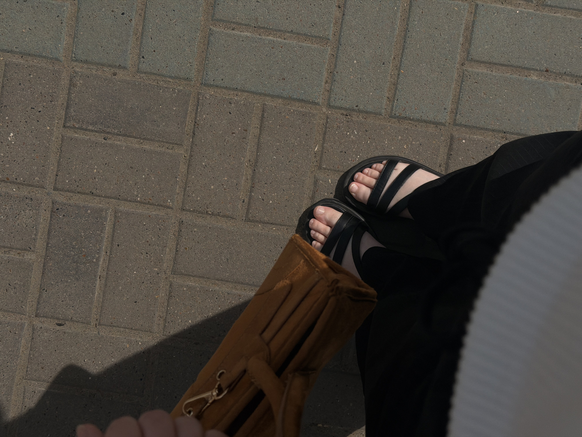 The image shows a view of the ground from above, likely taken by a person looking down at their feet. The ground is paved with gray rectangular bricks. The person is wearing black sandals with straps and black trousers. They are also holding a brown suede bag with a gold clasp. The location is presumed to be Chaikovsky, Russia. The subject is a person standing or sitting with their feet on the paved surface.