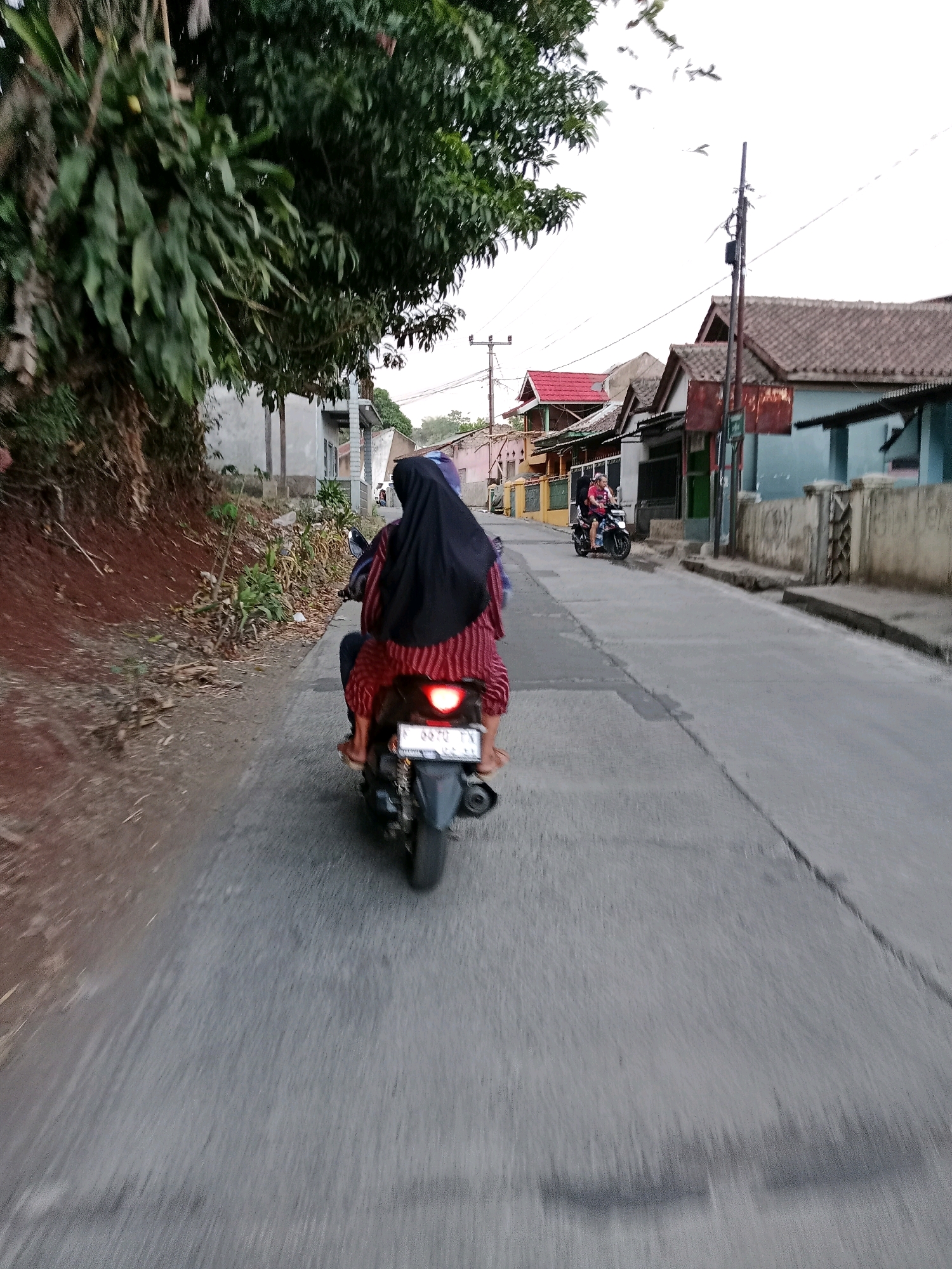 The image captures a residential street in Sirnagalih, Indonesia. In the foreground, two individuals are riding a motorcycle. The person in the back, wearing a black hijab and a red and white striped shirt, is the primary subject. The motorcycle's license plate is partially visible, reading "B 3570 TK". The road surface is asphalt with some cracks and debris. To the left, a steep, reddish-brown dirt embankment is covered with dry leaves and sparse vegetation. To the right, a row of houses with tiled roofs line the street. The houses are varied in color and architectural style, with some featuring fences and open doorways. In the distance, another person is visible riding a motorcycle. The sky is overcast, and there are a few birds in flight. The overall scene depicts a typical day on a local street in an Indonesian village.