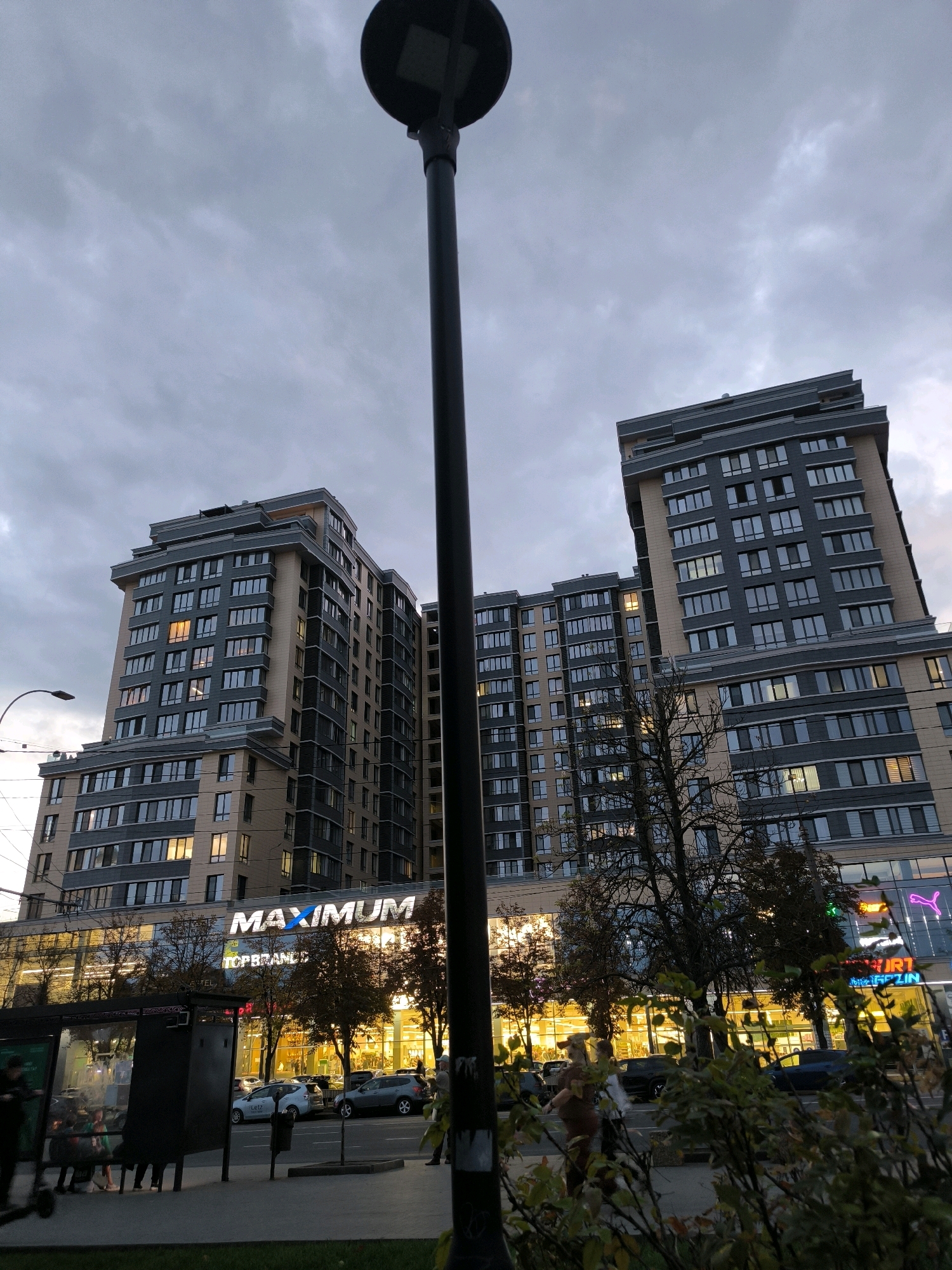This image captures a street scene in Chișinău, Moldova, at dusk. A large, modern apartment building with a retail space on the ground floor dominates the background. The building features a beige and grey facade with numerous windows, many of which are illuminated. The retail space below showcases a bright yellow interior and signage that includes "MAXIMUM" in large white letters and "TOP BRANDS". To the right, neon signs, including a pink Puma logo, are visible. A tall, dark lamppost stands prominently in the foreground, slightly off-center. In front of the building, trees with sparse autumn foliage line the street. A bus stop shelter with people sitting inside is situated on the left side of the image. Cars are parked and driving on the road in front of the retail stores. The sky is overcast with a cloudy, grey hue, indicating the end of the day.