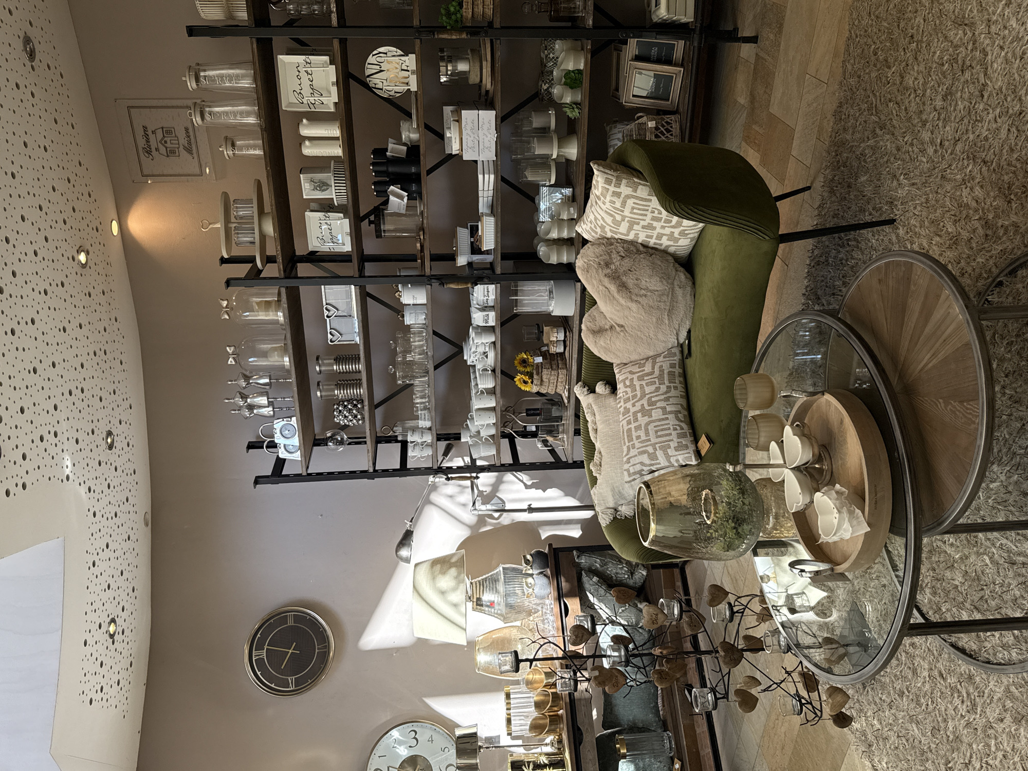 The image depicts the interior of a shop in Bad Sachsa, Germany, likely a home decor or gift shop. The scene is warmly lit by natural light streaming from the right, casting distinct shadows across the room. Dominating the foreground are two nested, round coffee tables with glass tops and dark metal frames. The larger table holds a decorative glass vase with green foliage inside, a wooden serving tray with four small white teacups and saucers, and a small white paper napkin. The smaller table is partially visible beneath the larger one, showing its wooden top.
To the right of the tables, a plush, olive-green sofa is arranged, adorned with several decorative pillows. Among them is a large, plush heart-shaped pillow and two patterned rectangular pillows. Behind the sofa, a tall, dark metal shelving unit is filled with a variety of items, including glassware, jars, decorative boxes, kitchenware, and framed pictures. The shelves are organized with items displayed in a retail manner.
To the left of the sofa and tables, a console table is visible, topped with several decorative items including lamps, vases, and a metal candle holder tree adorned with small wooden hearts. A large round clock with a dark face and Roman numerals is mounted on the wall behind the console table. The ceiling features a decorative pattern of small, scattered circles, with recessed lighting fixtures. The floor is a combination of tiled and carpeted surfaces. The overall ambiance suggests a well-arranged retail space showcasing home furnishings and decorative objects.