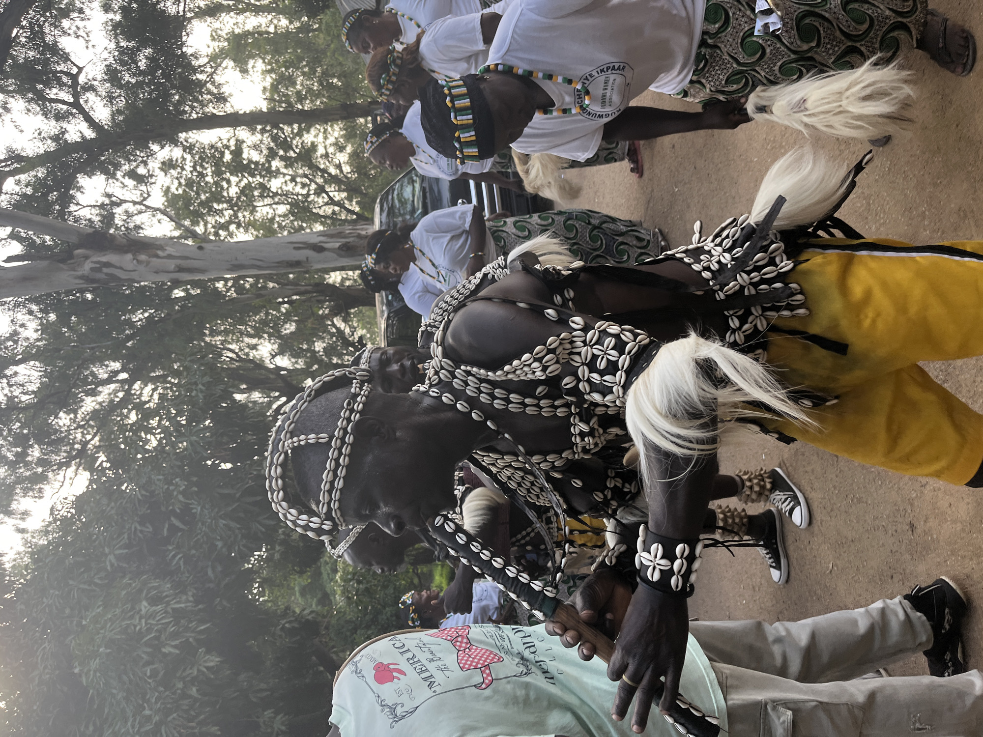 In a daytime outdoor scene on a dirt ground surrounded by trees, a group of people, predominantly women wearing matching white tops and patterned skirts, stand together. The central focus is a man adorned in traditional attire consisting of a headdress, vest, and bracelets, all decorated with cowrie shells. He is holding and playing a wooden flute. Another man, dressed similarly with cowrie shell decorations, stands beside him, and several other individuals are visible in the background, some holding fluffy white objects. The overall atmosphere appears to be a cultural gathering or ceremony, possibly in Jos, Nigeria, given the context.