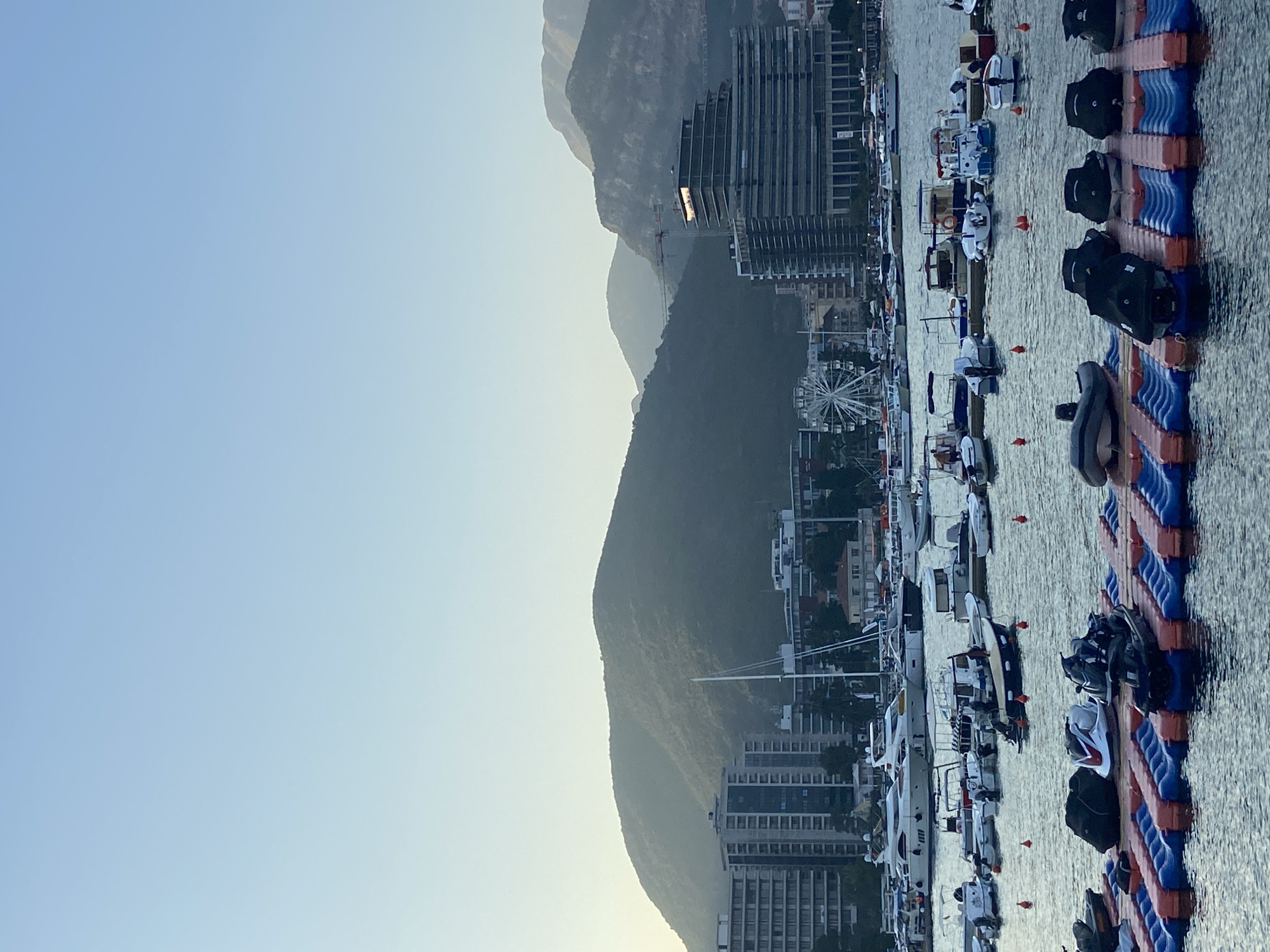 The image depicts a harbor scene in an urban area, with a clear sky and mountains in the background. The foreground features a floating dock system holding several jet skis. In the middle ground, numerous boats of varying sizes, including yachts and smaller motorboats, are docked in the water. Behind the boats, a cityscape is visible with multiple tall buildings and a Ferris wheel. The mountains behind the city are covered in lush greenery and appear to be illuminated by the soft light of dawn or dusk. The water is calm, reflecting the boats and surrounding structures.