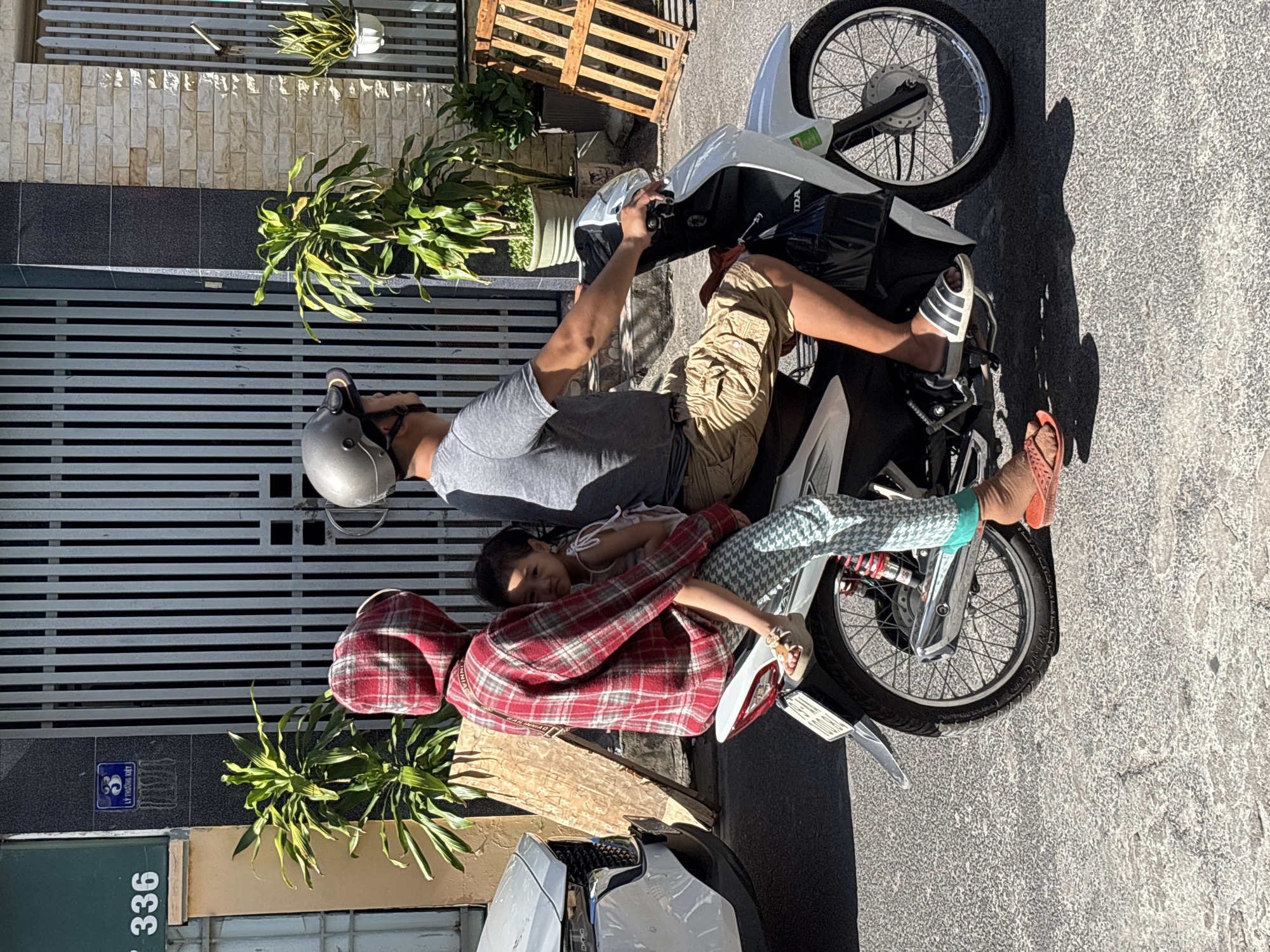 The image captures a street scene in Nha Trang, Vietnam, focusing on a family riding a white motorcycle. A man, wearing a grey t-shirt and khaki shorts, is driving the motorcycle, sporting a silver helmet. Seated behind him is a woman in a red and white plaid hooded jacket and patterned pants, holding a young child. The child, wearing a light-colored top, is cradled in the woman's arms and looks towards the camera.
The environment suggests a commercial or residential area with a building facade featuring a dark, textured wall and a light stone-patterned section to the right. A tall, barred gate is visible behind the subjects, flanked by lush green plants in pots. To the left, a white car is partially visible, and further left, a building with a green accent and the number "336" is noted. The ground is paved with asphalt. The overall impression is of everyday life in a Vietnamese city, with a focus on the intimate moment of a family traveling together.