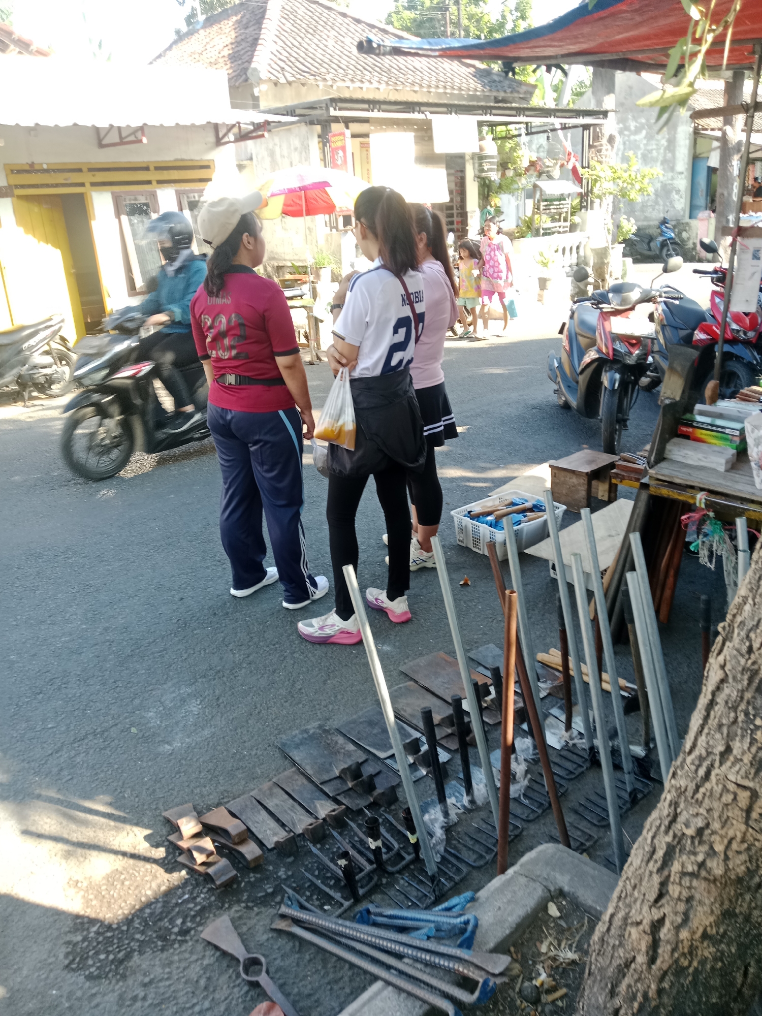 The image captures a street scene in the city of Kediri, Indonesia, on a sunny day. In the foreground, a collection of metal tools, including axes and rebar, are laid out on the asphalt. Behind the tools, three women stand on the street. One woman in the foreground wears a maroon shirt and navy pants, with a beige cap on her head. Another woman to her left wears a white shirt with blue lettering and a black skirt. The third woman is partially visible behind the second, wearing a pink shirt and yellow shorts. A motorcycle is visible to the left, with a person in a blue jacket riding it. In the background, there are buildings with tiled roofs and a red and blue awning. An umbrella with red and white stripes is also visible in the middle ground. The overall scene suggests a bustling street market or roadside stall.