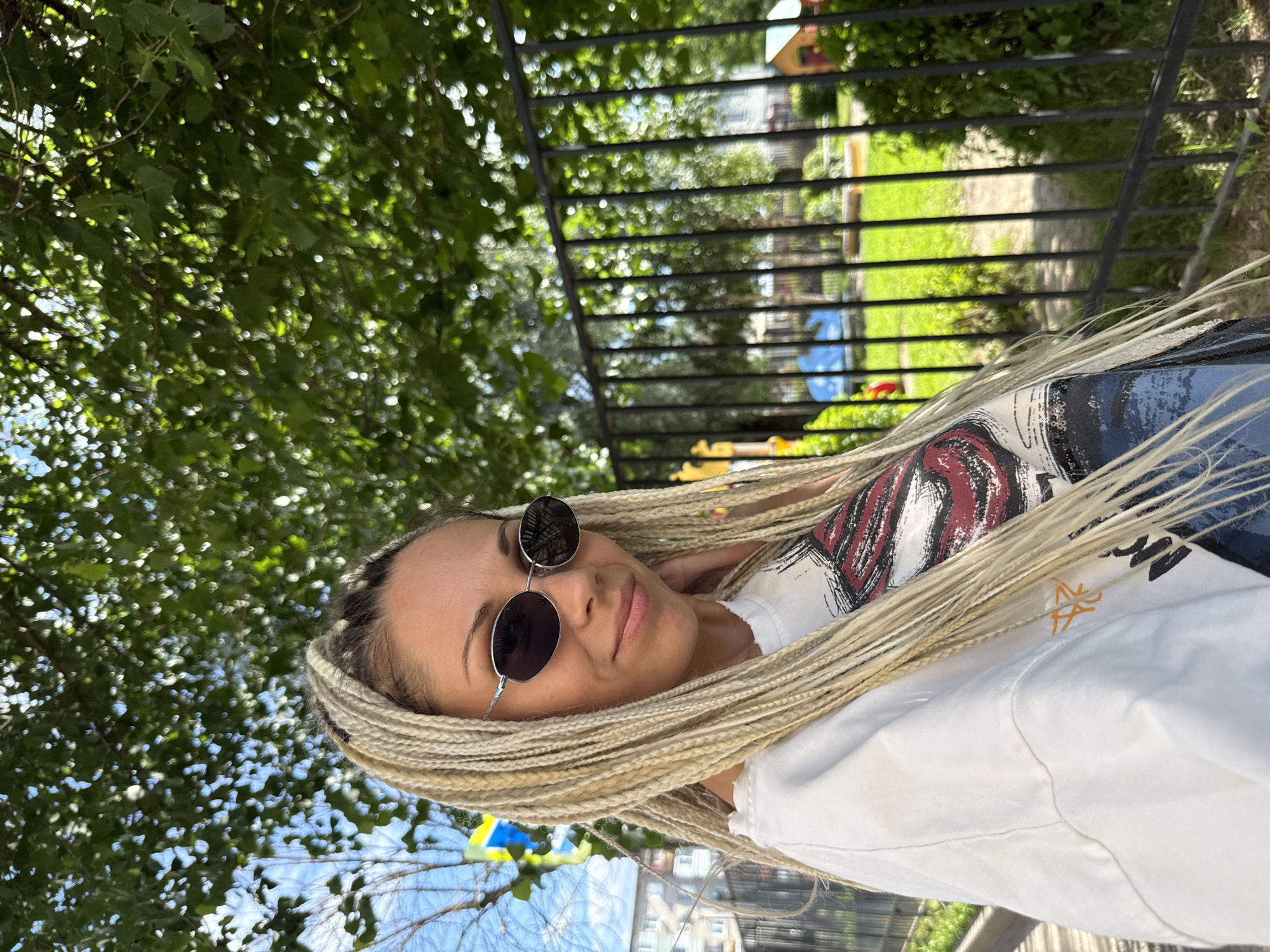 A woman with blonde braided hair and sunglasses is posing for a photo outdoors. She is wearing a white t-shirt with a graphic print and blue jeans. Behind her, there is a black metal fence, green trees, and a playground with colorful equipment. The scene appears to be a residential area in Rusanovo, Russia, given the presence of apartment buildings in the background and the overall suburban aesthetic. The lighting suggests it is daytime, with sunlight filtering through the leaves of the trees.