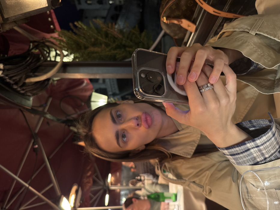 An adult female is depicted in an outdoor restaurant setting at night in Venice, Italy, with the Rialto Bridge visible in the background. She is centrally framed, looking towards the viewer with a "duck face" expression, lips puckered. She holds a gray smartphone, identified as an iPhone Pro model in a clear MagSafe-compatible case, with both hands, positioned in front of her chest. Her left ring finger displays two rings: a simple silver band and a diamond eternity band. Her nails are manicured with a clear or light pink polish. She wears a beige trench coat over a blue and white plaid shirt, the cuff of which is visible on her left arm.
The environment includes red awnings or umbrellas overhead, fitted with active radiant heaters. An ornate, illuminated lamp hangs to the right. To the left, blurred figures of other diners and a table with a green bottle are visible. In the deep background, beyond a visible canal and green foliage, the illuminated, distinctive arch of the Rialto Bridge is clearly discernible, flanked by light-colored buildings. A brown Louis Vuitton monogrammed bag is partially visible on a surface to the right. The overall lighting suggests ambient illumination from restaurant fixtures and distant city lights.