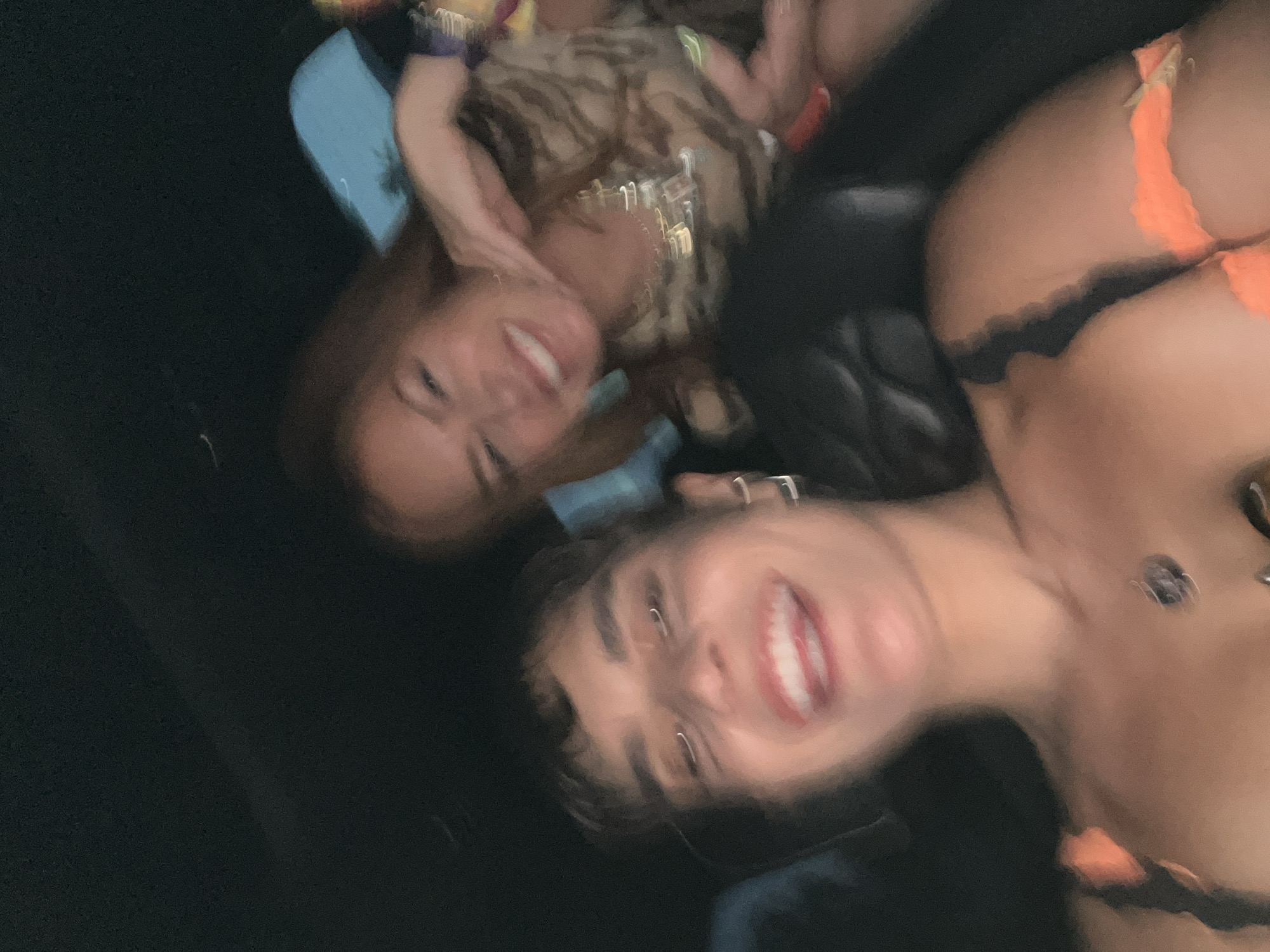 The image captures three individuals in what appears to be a dimly lit interior, possibly a vehicle. The primary subject in the foreground is a person with short, dark hair, fair skin, and a visible pendant necklace. They are smiling broadly, revealing their teeth. To their upper left is another individual with reddish-brown hair and fair skin, also smiling. The third person is partially visible in the background, wearing a patterned top and jewelry. All subjects appear to be in motion or the photograph was taken with camera movement causing blurriness. The location is unknown; there is no information that it is in La Quinta, United States of America.