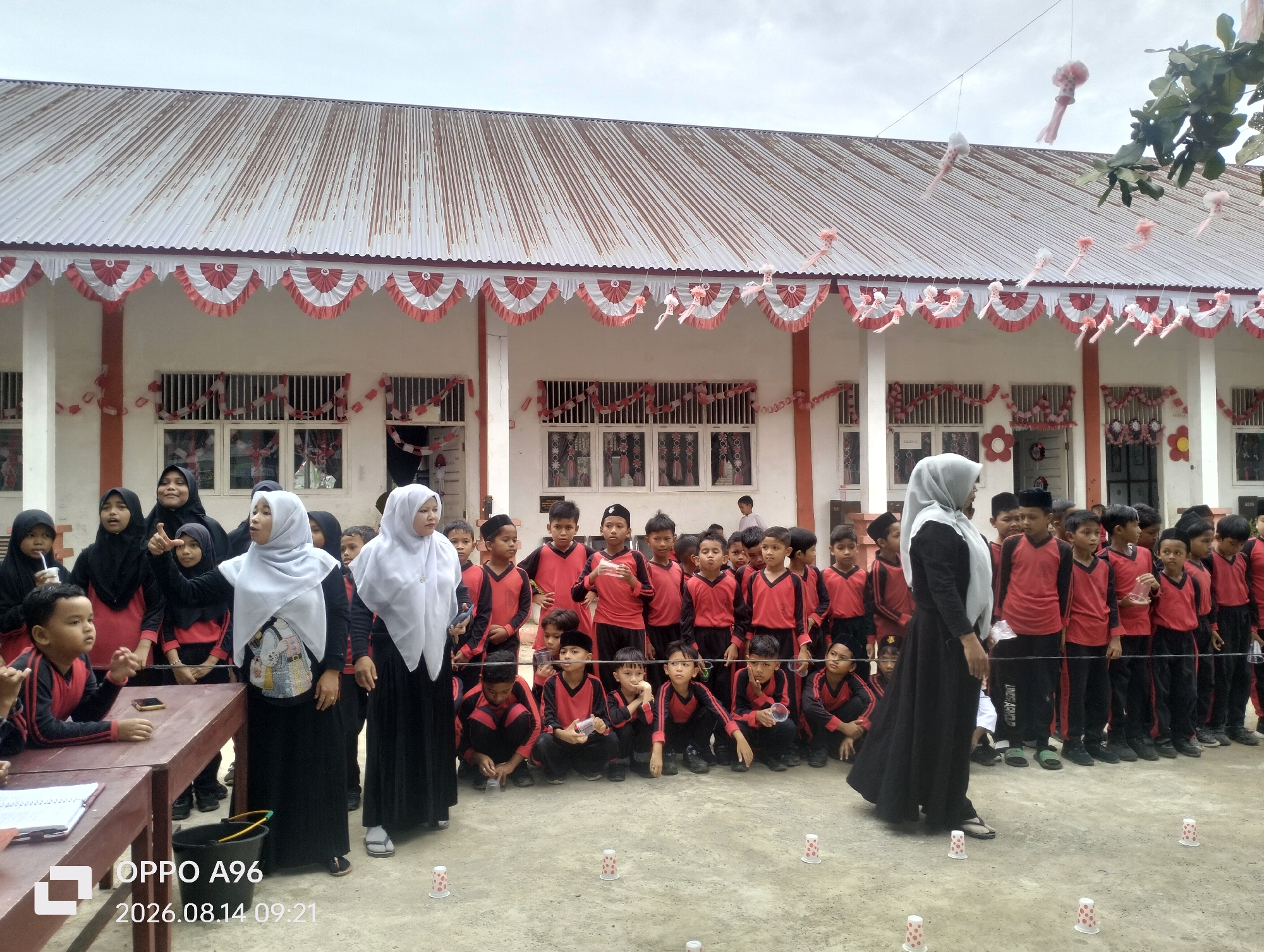 The image captures a group of children, likely students, gathered outdoors in front of a building with a corrugated metal roof. Many of the children are wearing matching red and black uniforms, and several of the adults present are wearing hijabs. The building's facade is decorated with red and white bunting and paper lanterns, suggesting a festive occasion. The ground is paved with concrete. In the foreground, there is a wooden table with papers and a phone on it, and a black bucket. Several small, polka-dotted cups are arranged in rows on the ground. The timestamp at the bottom of the image indicates the photo was taken on August 14, 2026, at 09:21. The setting is the grounds of a school, possibly in Keudekarieng, Indonesia, given the context.