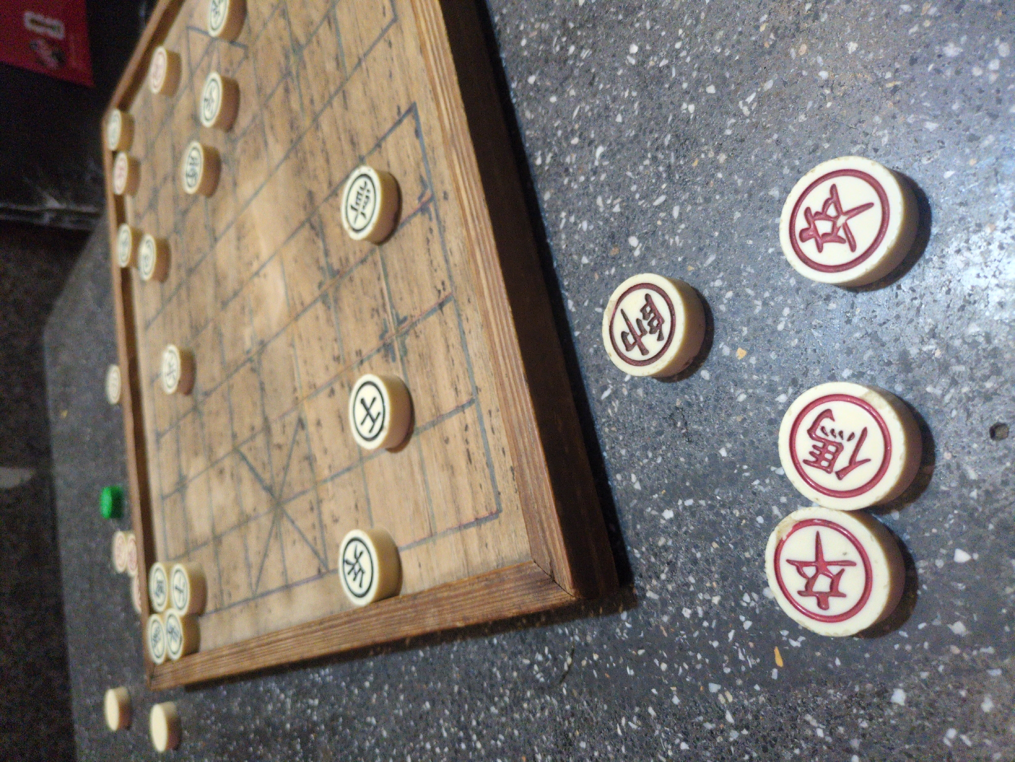 A wooden Xiangqi (Chinese Chess) board and several game pieces are positioned on a dark, speckled countertop. The rectangular board, crafted from light brown wood, displays a grid of darker lines, including the central "river" and diagonal lines within the "palace" areas. Approximately twenty-five circular, off-white game pieces with red Chinese characters are scattered across the board, suggesting a game in progress or recently concluded. An additional four identical pieces are arranged on the speckled countertop in the foreground, with one separate green cylindrical object visible near the upper left of the board. The countertop is dark gray or black with white and light gray speckles. The background is dark and out of focus. This scene is located in South Quy Nhon Ward, Vietnam.