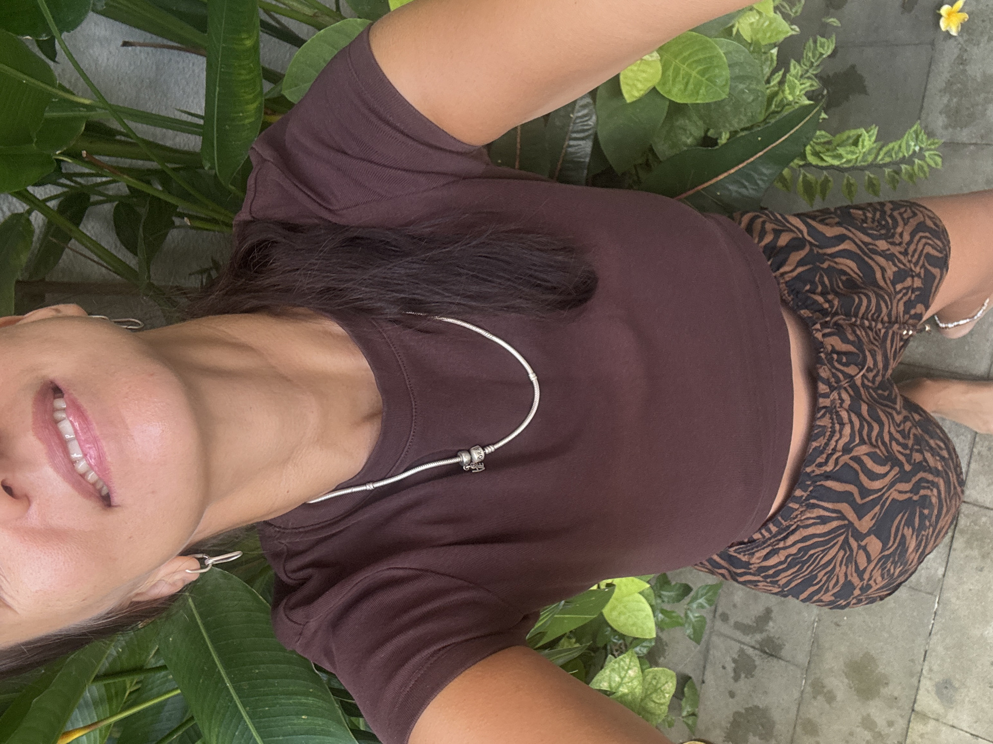 The image is a bright, daytime selfie taken by a person outdoors in what appears to be a tropical garden or patio setting in Kuta Selatan, Indonesia. The person's face is partially visible on the left, tilted slightly upwards and smiling with light pink lips and white teeth. They are wearing a plain brown short-sleeved t-shirt and patterned shorts featuring a black and brown animal print, possibly tiger or zebra stripes. A silver chain necklace with a charm, faintly displaying the text "PANDORA", is visible on their chest. Silver hoop earrings adorn their ears, and a silver anklet is visible on their right ankle. The background consists of dense, lush green foliage and grey paved ground. A single small yellow flower rests on the ground in the upper right corner.