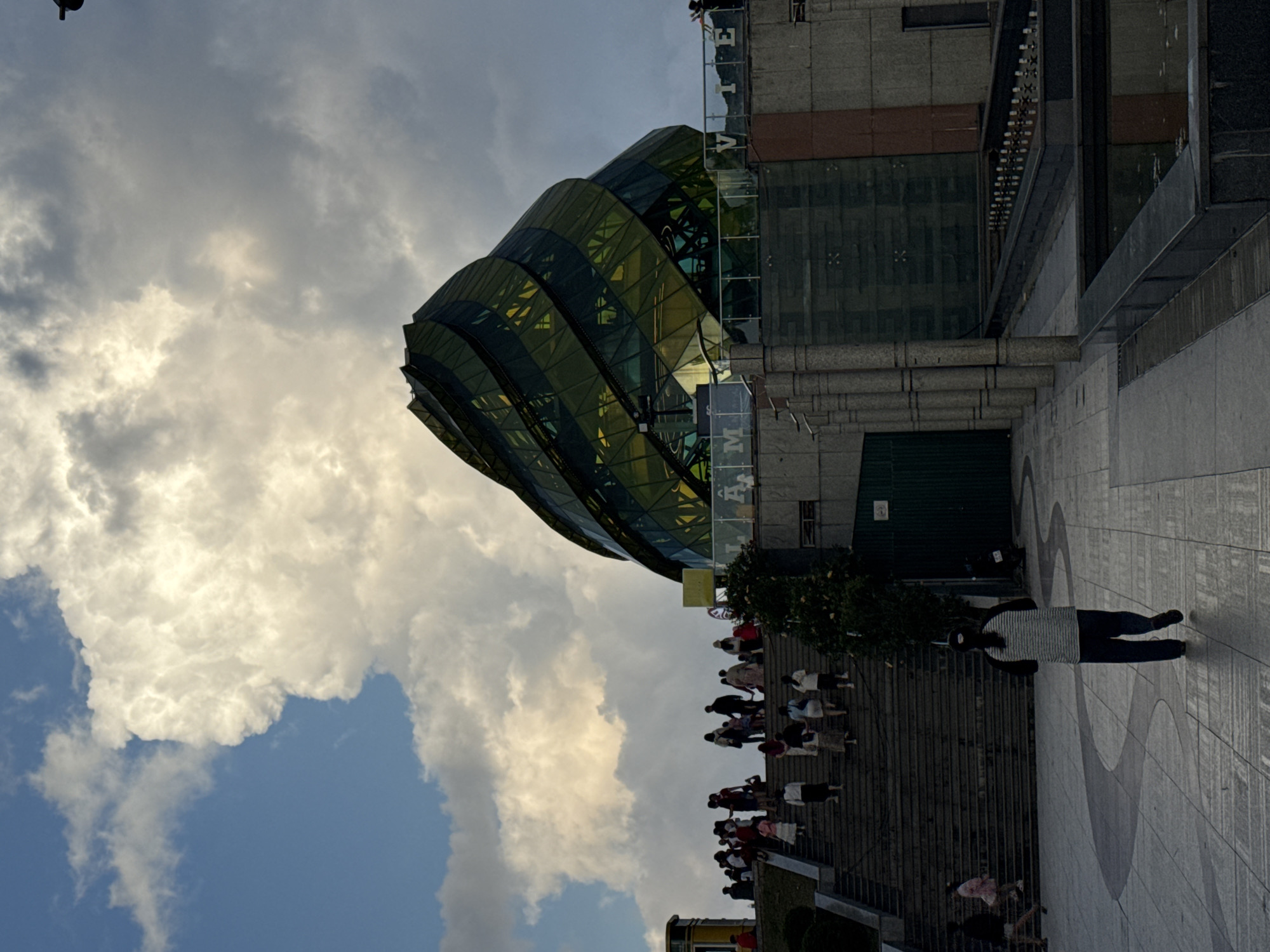 The image captures a scene at the Da Lat Flower Garden in Da Lat, Vietnam, on a partly cloudy day. A unique, spiral-shaped building with green and yellow glass panels dominates the background. In the mid-ground, a wide set of concrete stairs is filled with people walking up and down, many silhouetted against the bright sky. A lone figure walks away from the camera on a patterned paved walkway in the foreground. To the right, a long, dark stone structure with water features is visible. The overall atmosphere suggests a popular tourist destination with a blend of natural elements and modern architecture.
