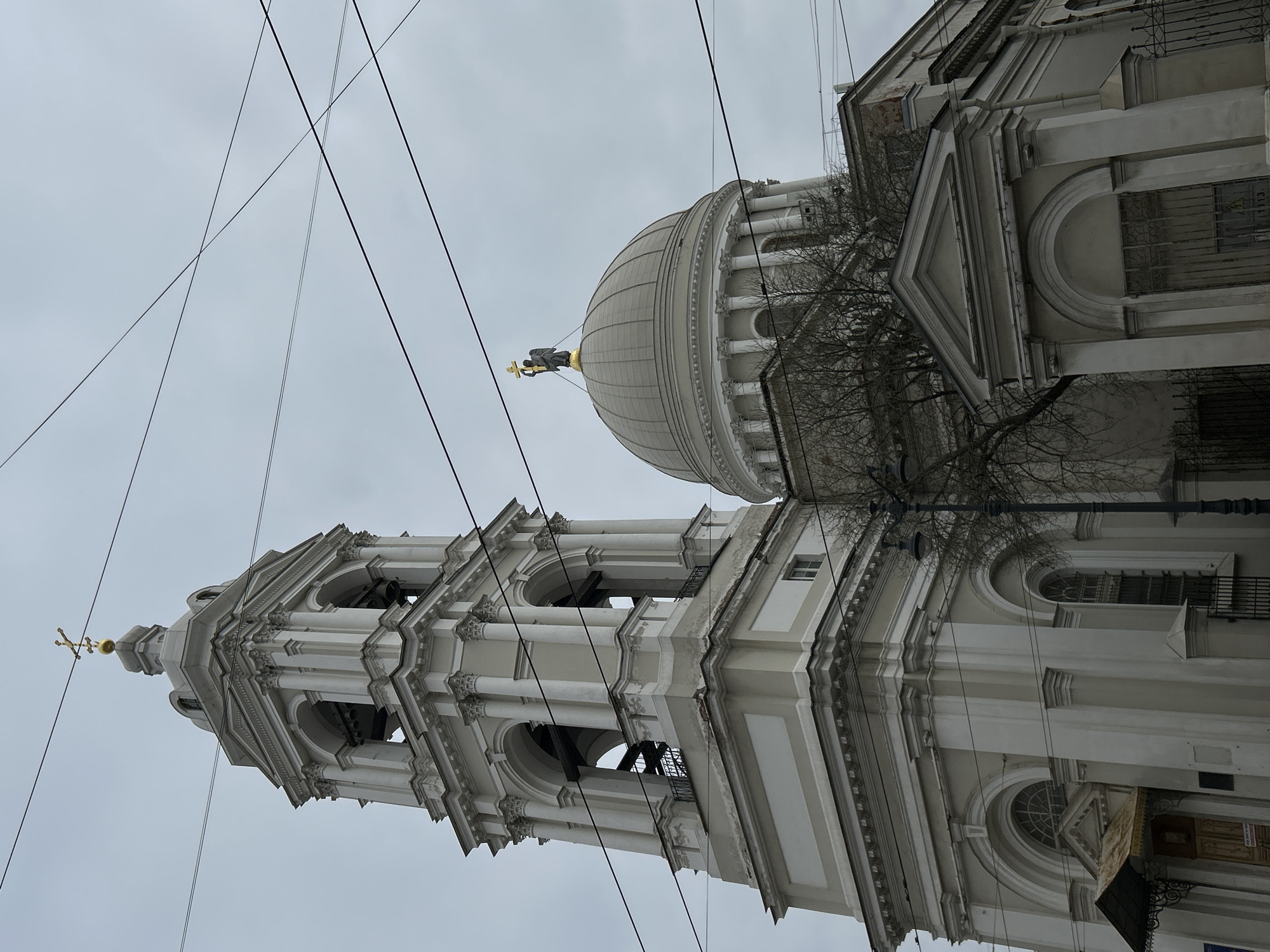 This image presents a grand architectural edifice, likely a church or cathedral, under a uniform overcast sky. The photograph is taken from a low angle, looking up at the structure, and is oriented vertically, displaying the buildings sideways in the original file.
The main subjects are a tall, multi-tiered bell tower and a prominent dome, both constructed from light-colored, possibly off-white or light grey, stone or stucco. The bell tower features successive levels of arched openings supported by classical columns and is topped with a golden cross. Adjacent to the bell tower, a large hemispherical dome rises, encircled at its base by a colonnade. A golden statue, appearing to be an angel holding a cross, crowns the dome.
The architectural style is classical or neoclassical, characterized by intricate detailing such as cornices, pilasters, and decorative moldings visible on the building's facade. Arched windows, some with protective grilles, are also discernible. Bare tree branches, indicative of winter or early spring, are scattered across the scene, partially obscuring sections of the building.
The sky is a muted, light grey, suggesting a cloudy day without direct sunlight. Numerous dark utility wires or power lines crisscross the upper portion of the frame, traversing the sky and the buildings. No people are visible, and no specific activity is taking place; the image is a static architectural portrait. The setting suggests an urban environment.
Faint, indistinct text or numbers, possibly "113" in Cyrillic script, can be seen on a building element in one of the cropped views, providing a subtle location cue within the given context of Saint Petersburg, Russia.