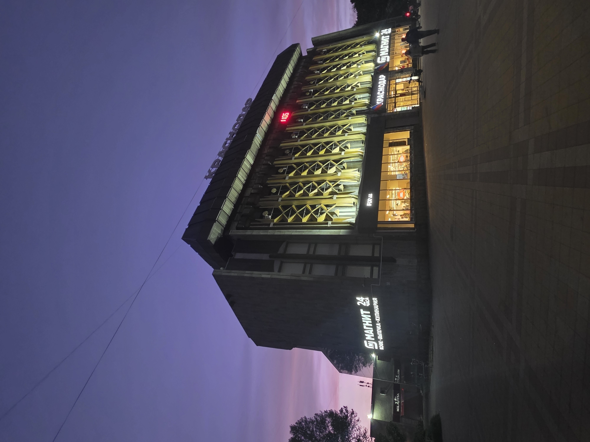 The image shows a multi-story building, likely a commercial complex, in Krasnodar, Russia at dusk or night. The building is labeled "Krasnodar" in Russian and has a "Magnit 24" sign, which is a popular convenience store chain in Russia. The building's facade has decorative elements. A digital clock displays the time "1:05." People are visible near the building entrance. The sky is a mix of purple and blue. The environment is urban.